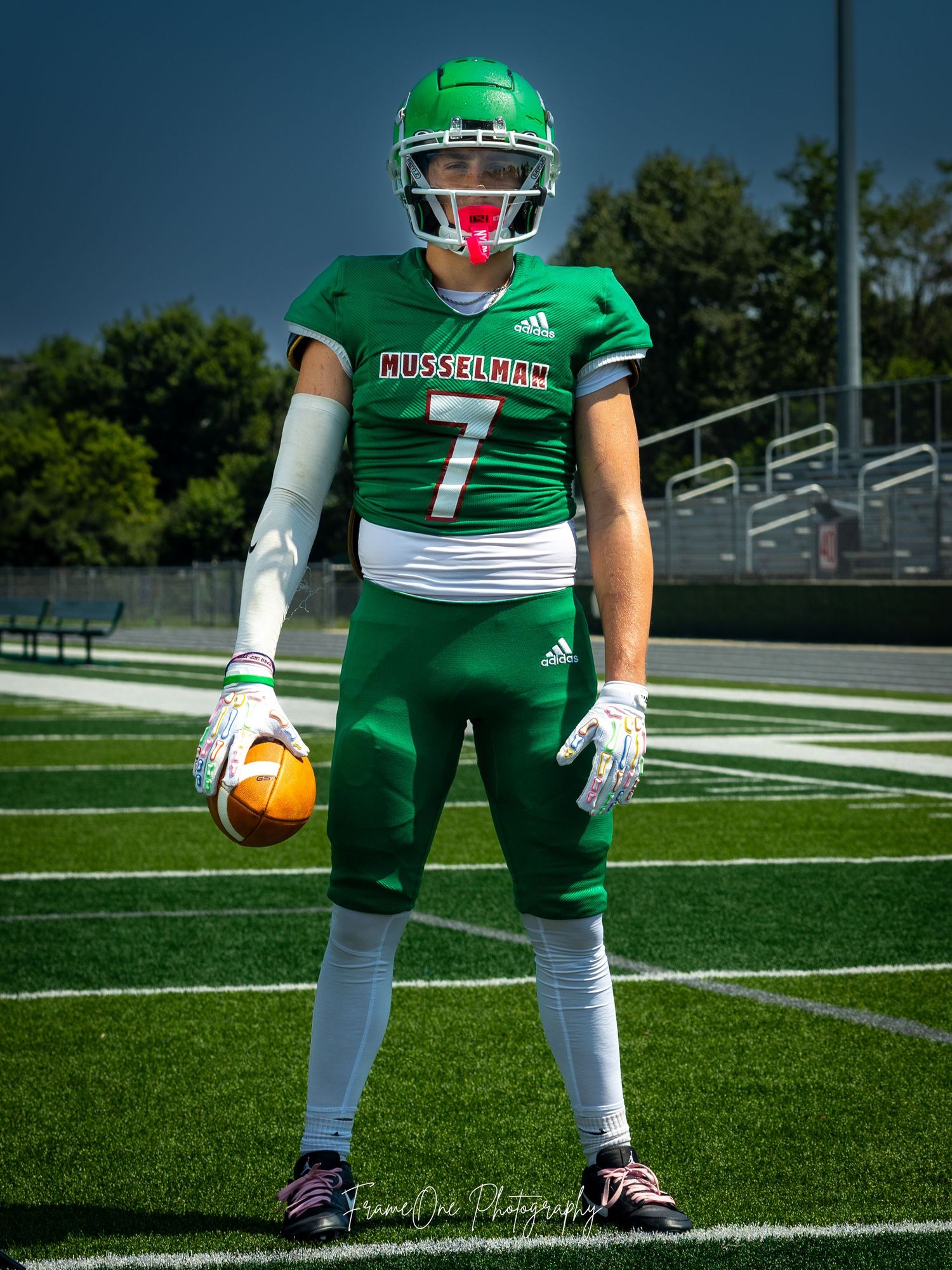 Football player in green uniform with helmet and number 7 jersey stands on field holding ball.