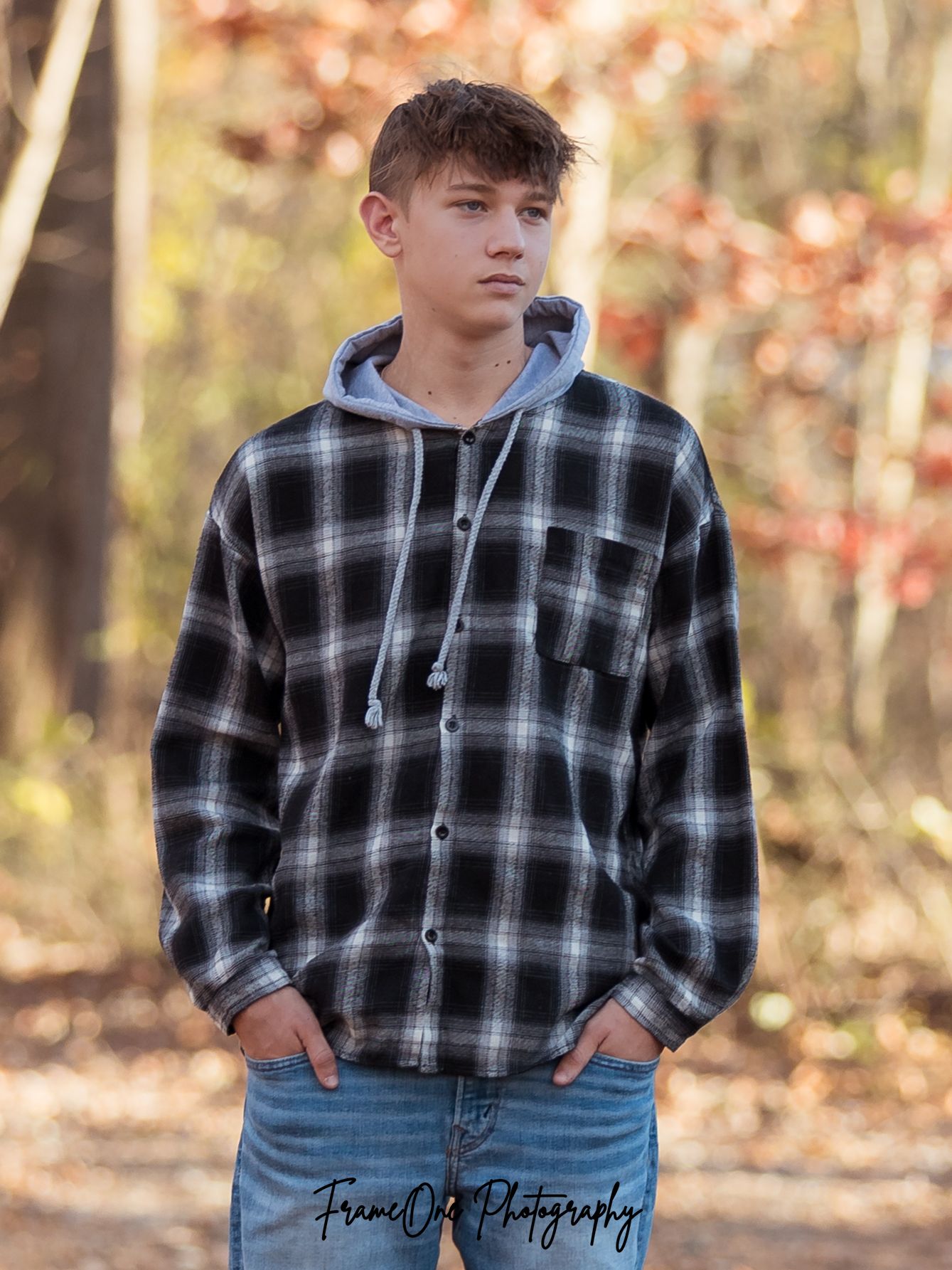 A person in a black and white plaid flannel shirt and jeans poses outdoors against an autumn forest background.