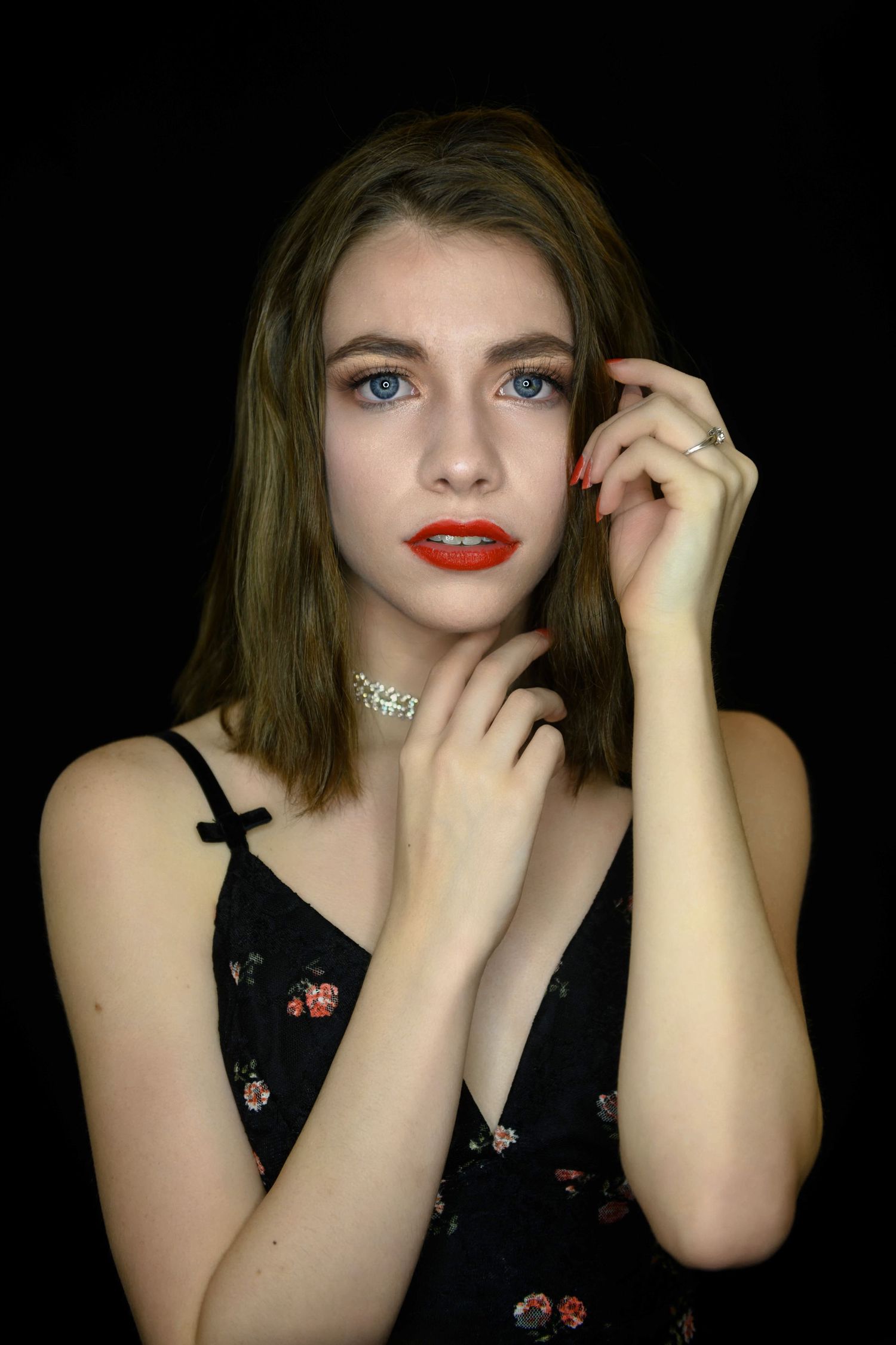 Portrait with red lipstick and floral black dress against dark background, wearing delicate necklace.
