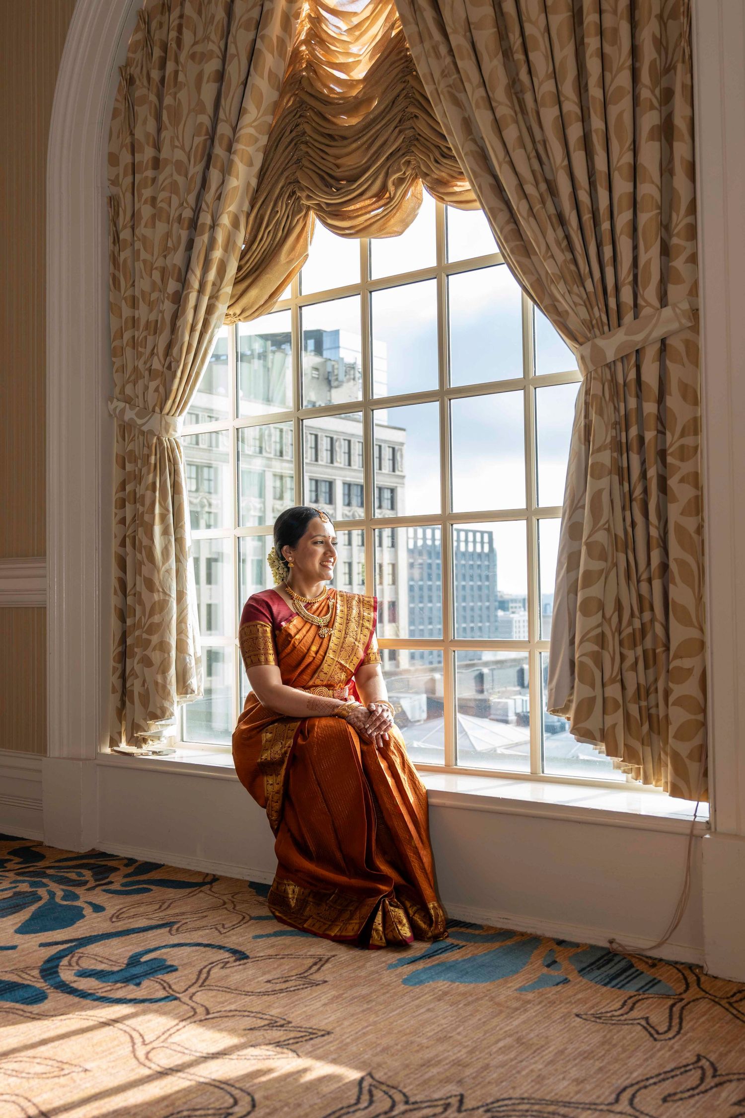Figure in traditional orange sari sits elegantly by ornate window with draped curtains overlooking city buildings.
