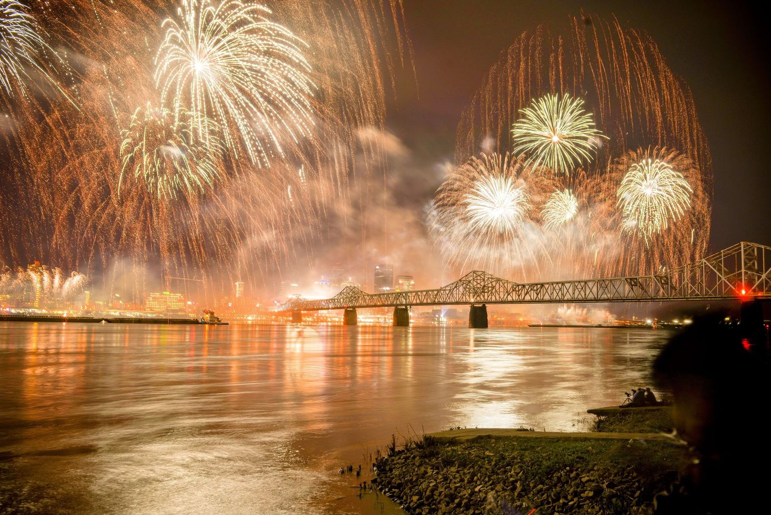 Spectacular fireworks display illuminates a bridge and river at night with golden bursts.