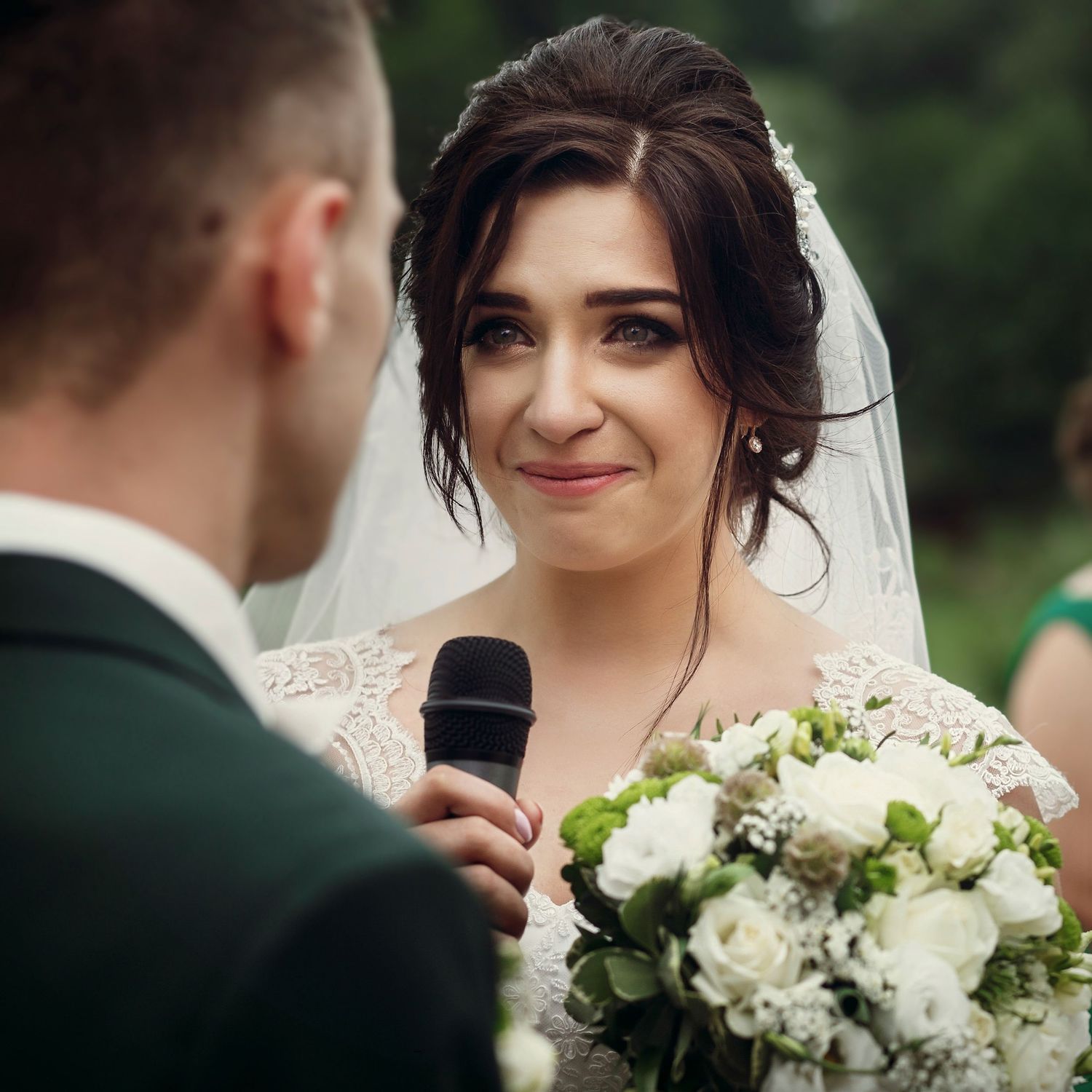 Bride in white dress holding bouquet smiles emotionally while groom speaks into microphone during ceremony.