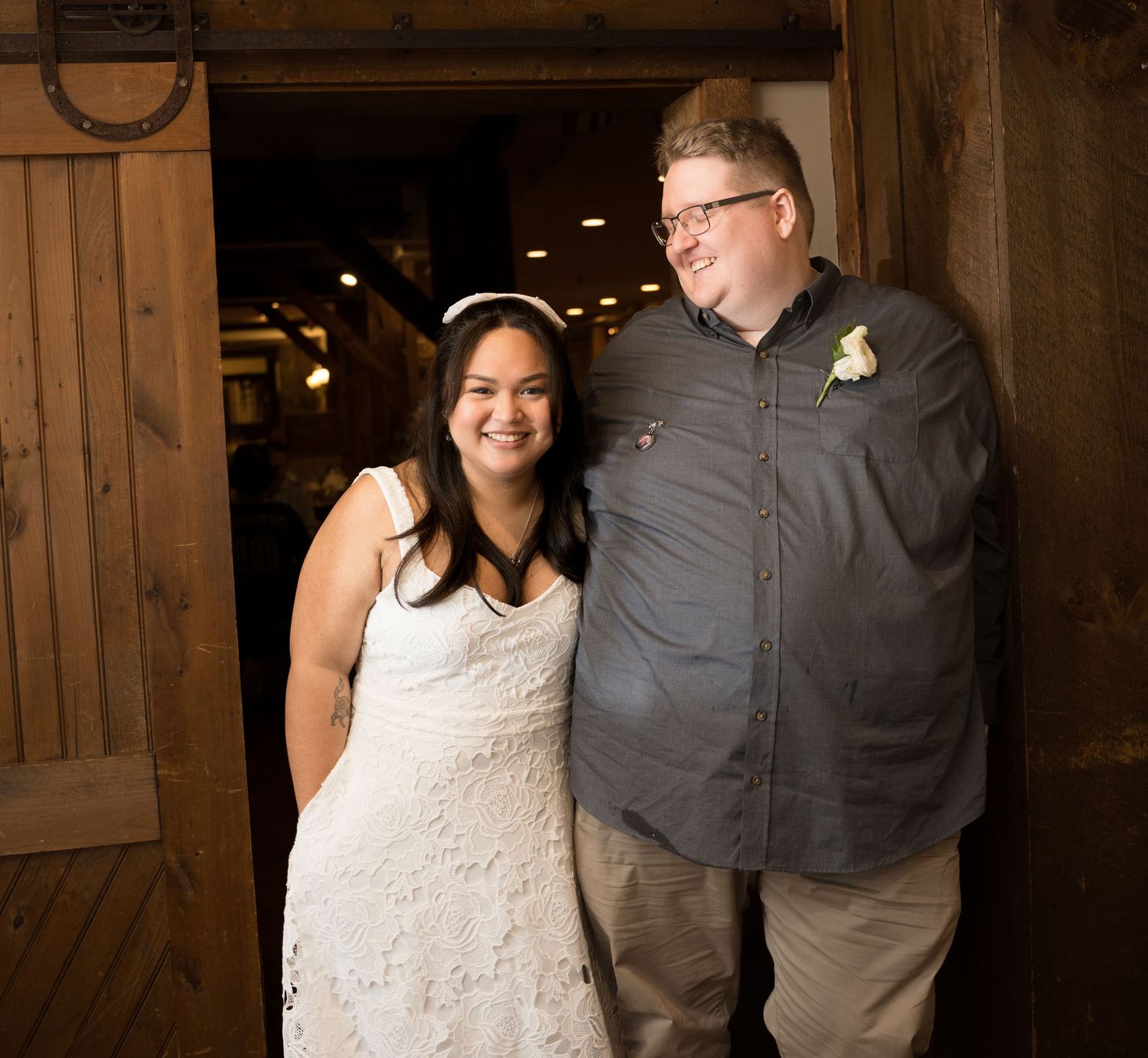 A happy couple smiles while standing in a rustic barn venue, with the bride wearing a white lace dress.