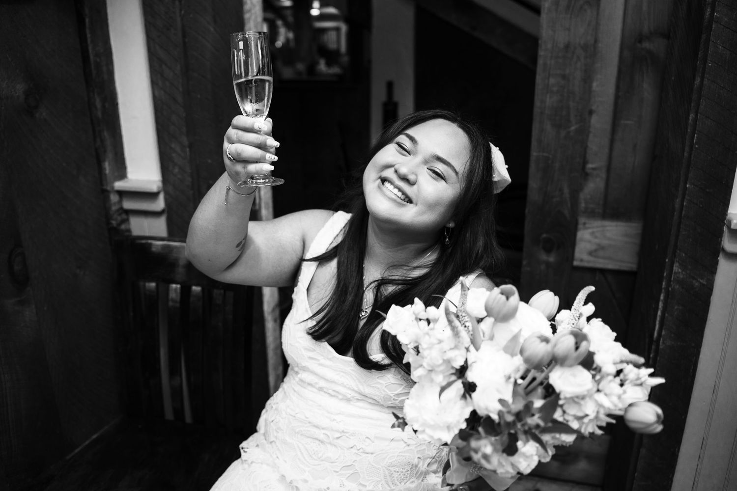 Woman in white dress raises champagne glass with white flowers sitting on a chair at reception.