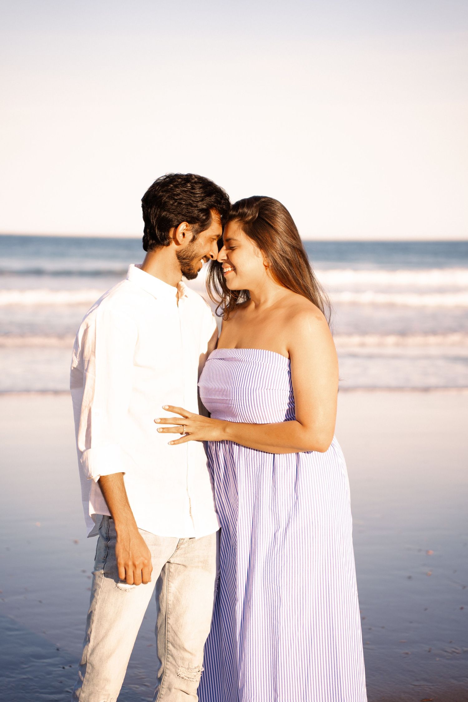 A romantic couple in light colored clothing share an intimate moment together on a serene beach at sunset.