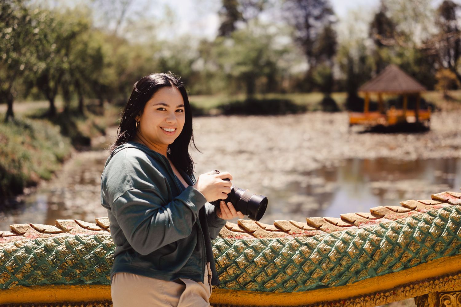 Photographer holding camera on ornate yellow bridge railing with scenic park pond in background during autumn.