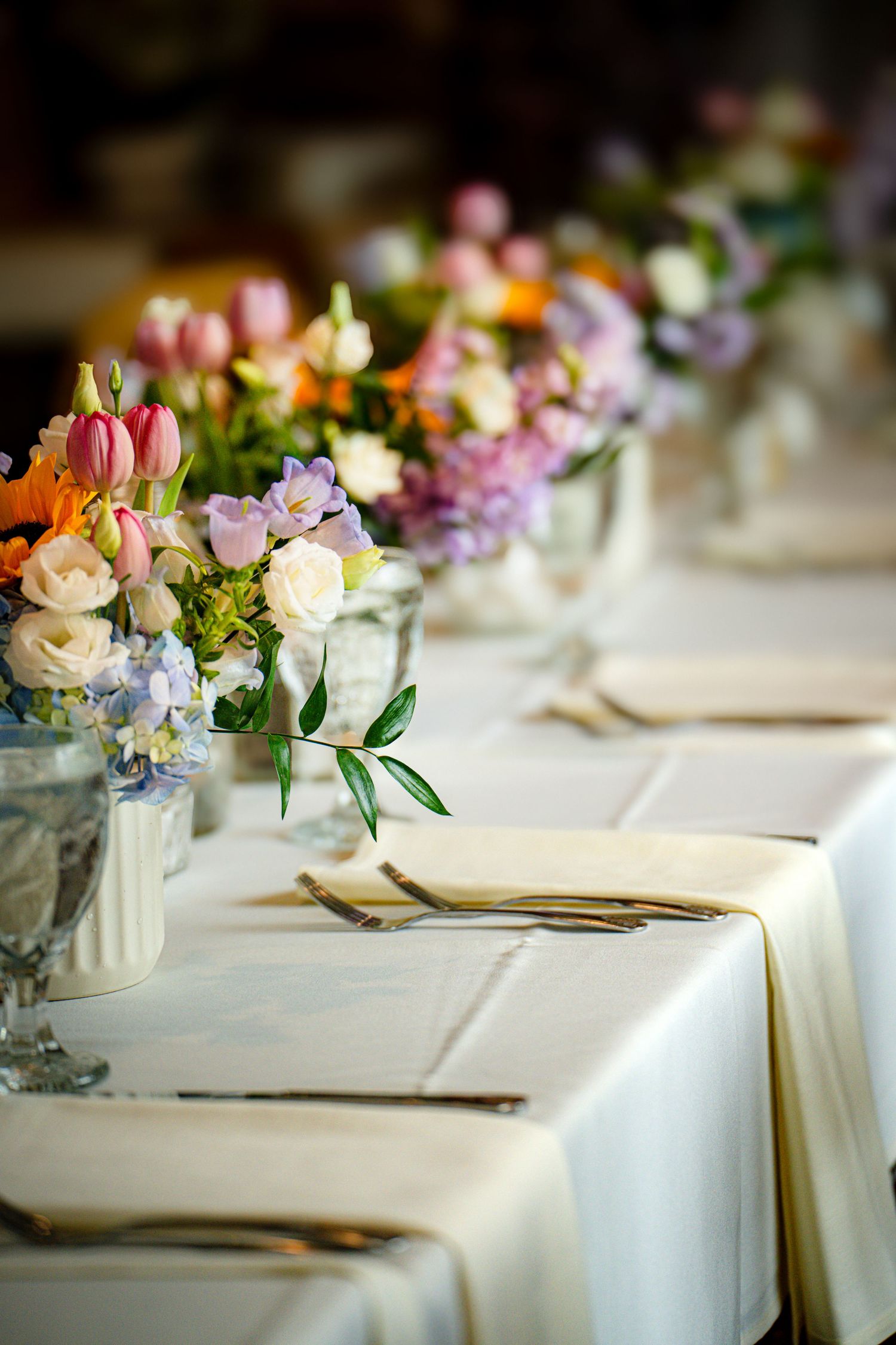 Elegant table setting with colorful spring floral centerpieces in glass vases on white linen with golden cutlery.