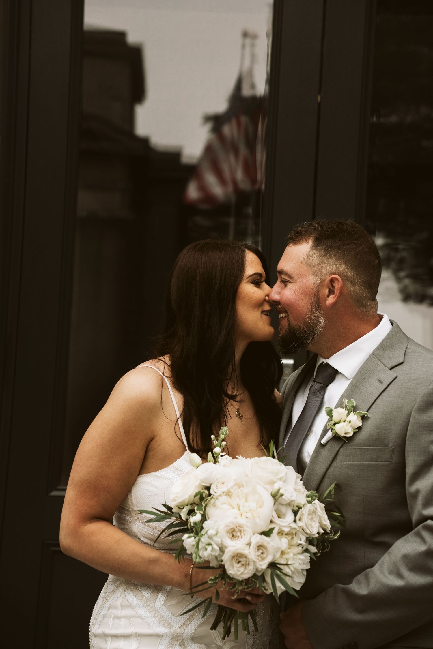 Romantic wedding couple sharing an intimate moment while holding a white rose bouquet with American flag in background.