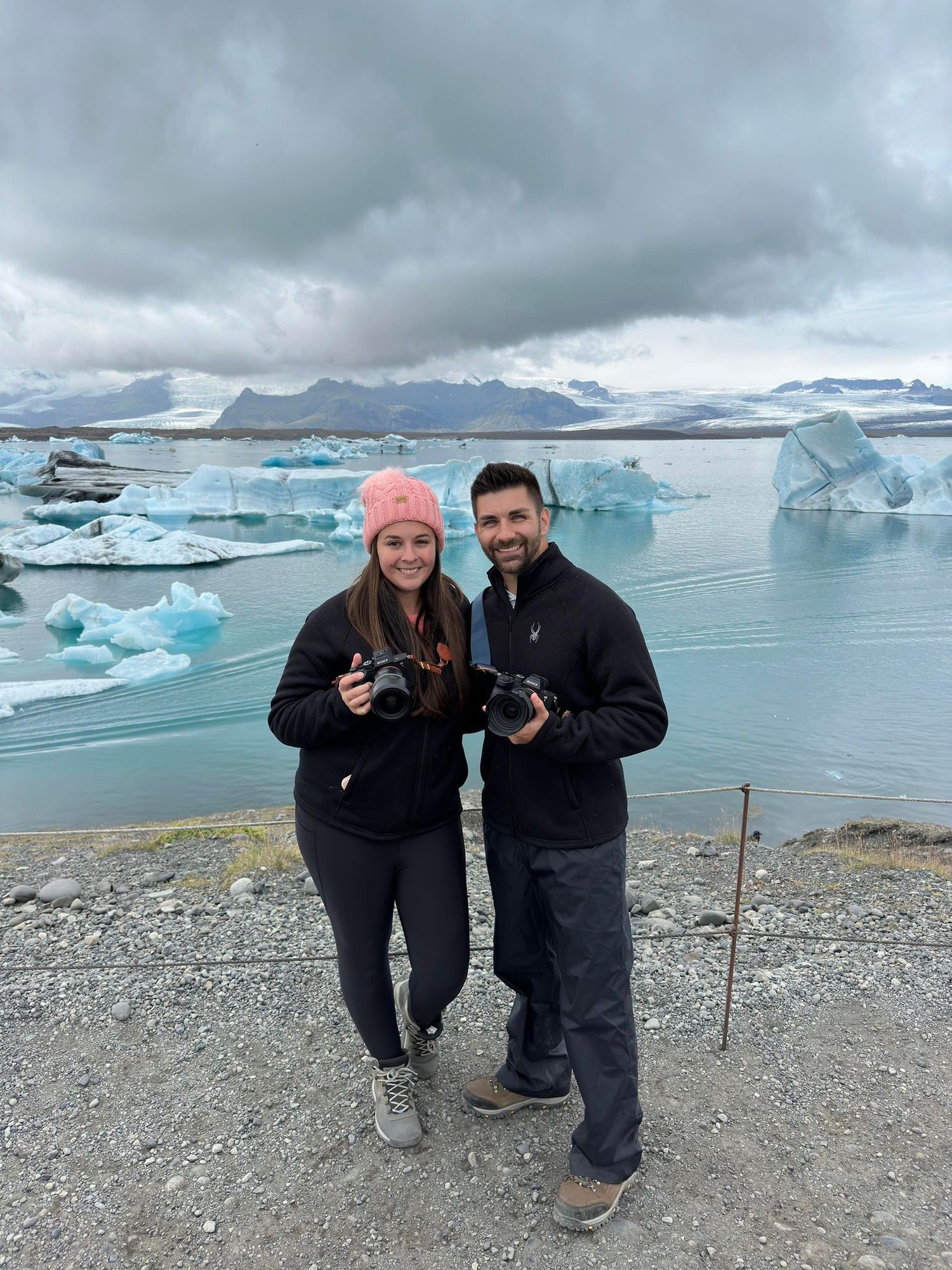 Two photographers stand along an icy shore with glacial lagoon and mountains in the background in cloudy Iceland.