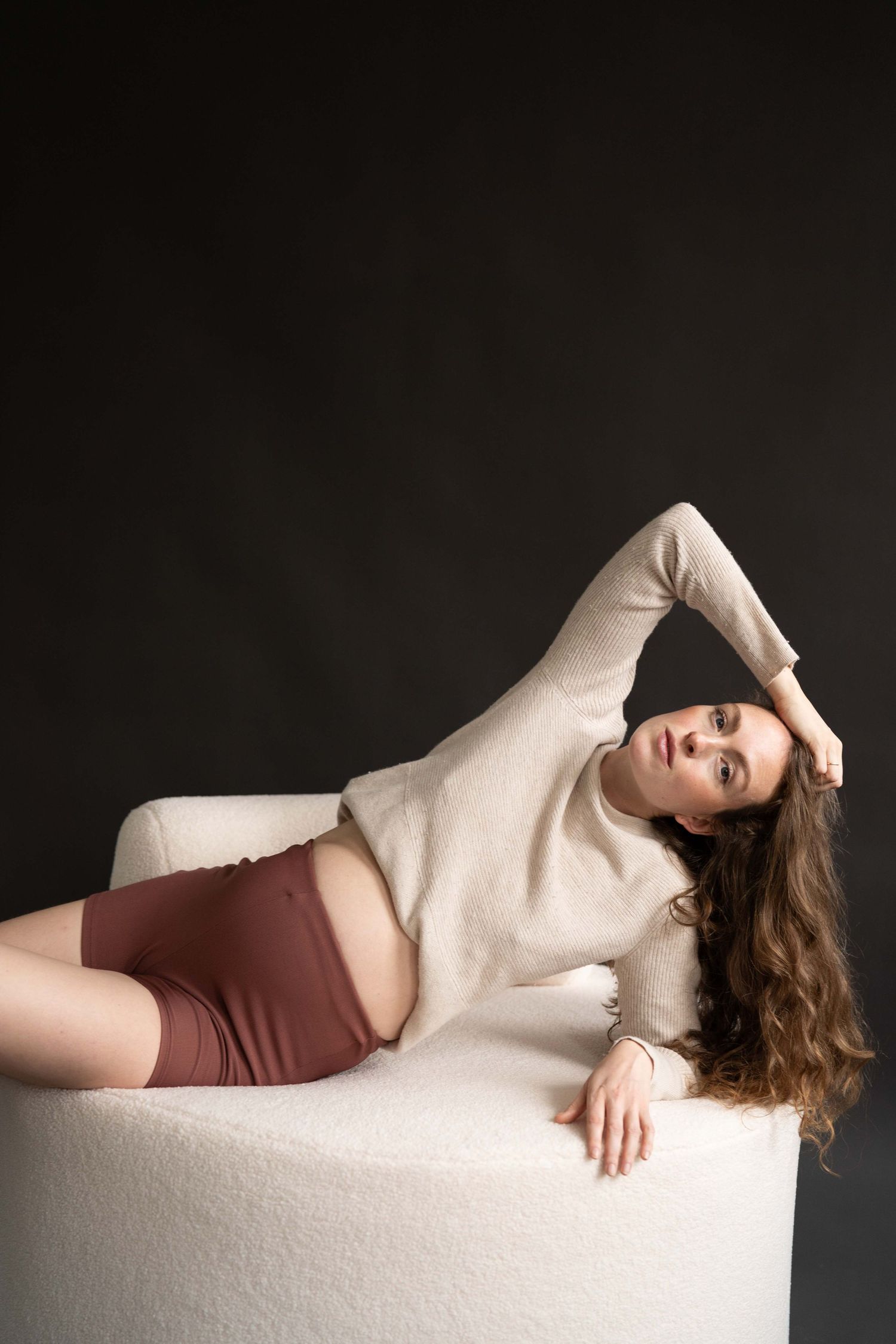 Schwangerschaftsshooting Berlin Studio – schwangere Frau in beigem Longsleeve und rostroter Shorts posiert elegant auf weißem Hocker