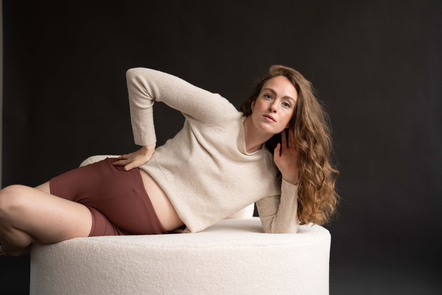 Babybauch Fotografie Berlin – schwangere Frau liegt lässig auf weißem Polsterhocker, modernes Maternity Shooting im Studio, The Elephant Lab