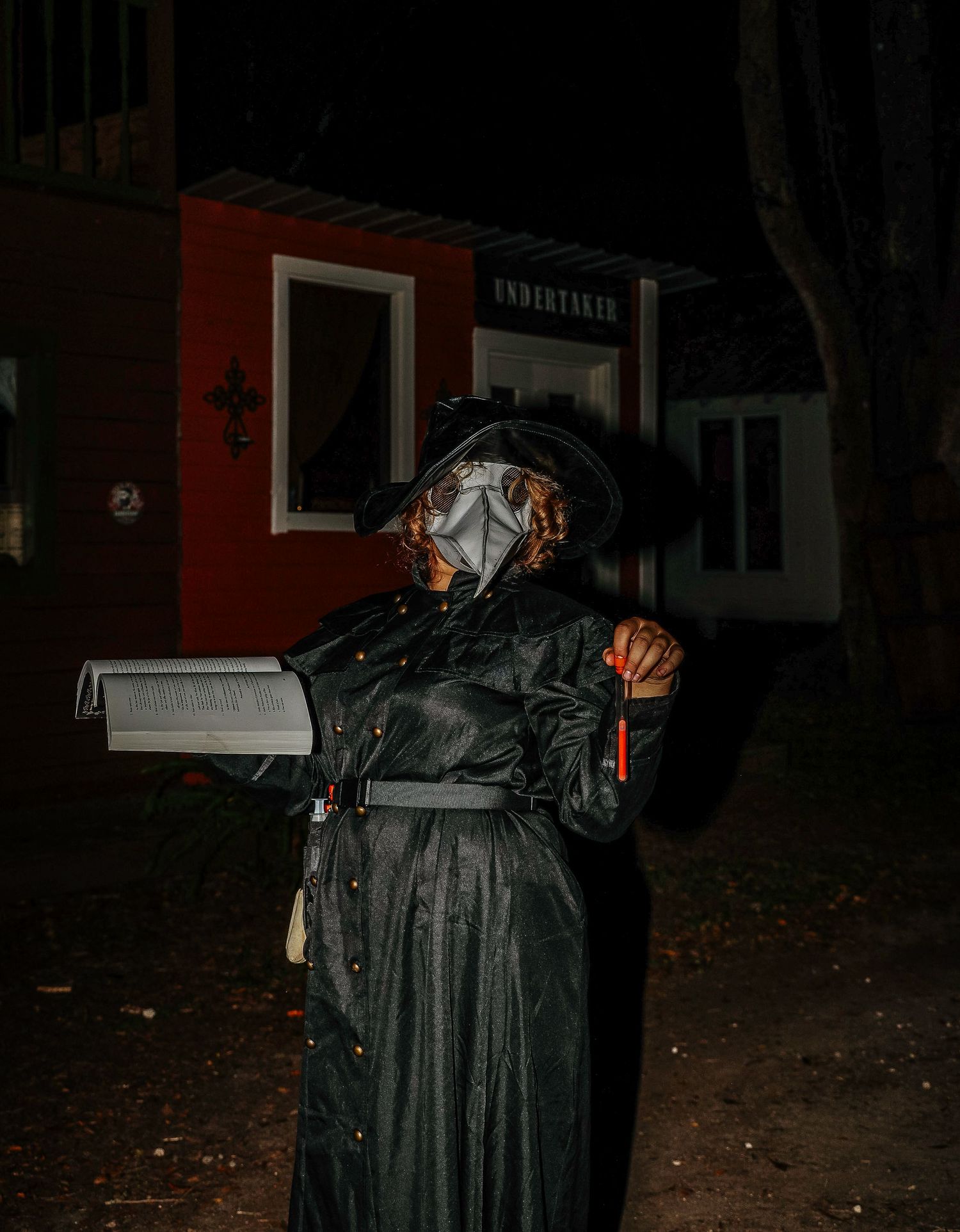 A person in a dark Halloween costume stands in front of a house at night holding a glowing pumpkin decoration.