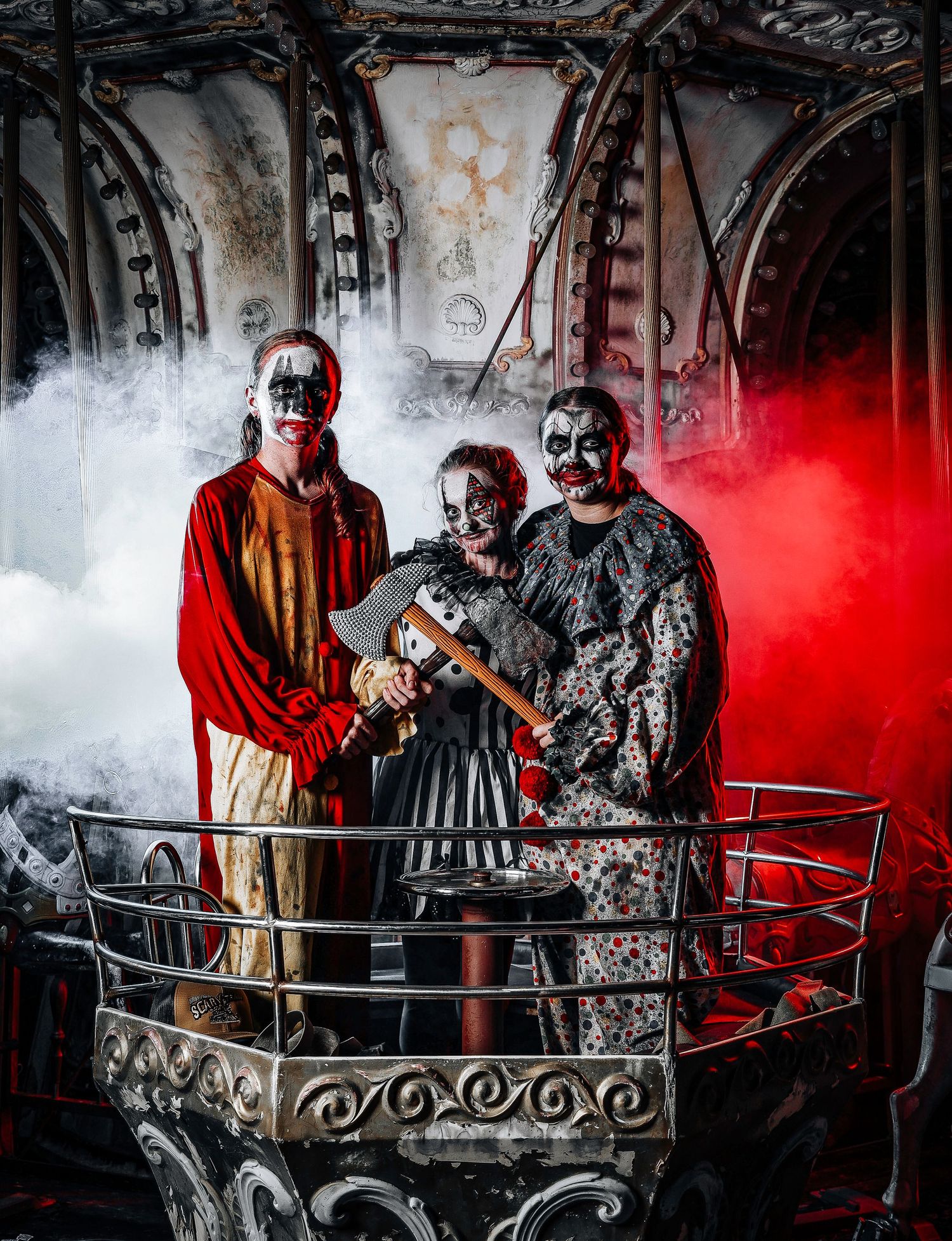 Three figures in face paint and gothic attire pose dramatically in fog with red lighting on a metal balcony structure.