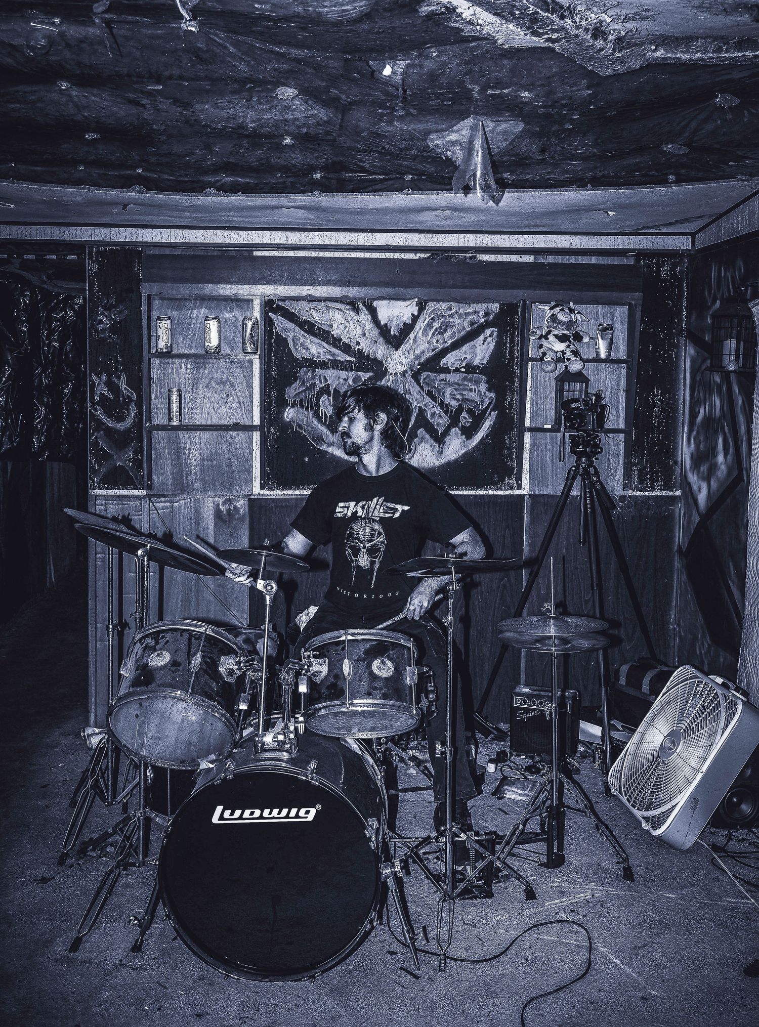 Black and white photo of a drum kit setup in a basement with band posters and equipment visible in the background.