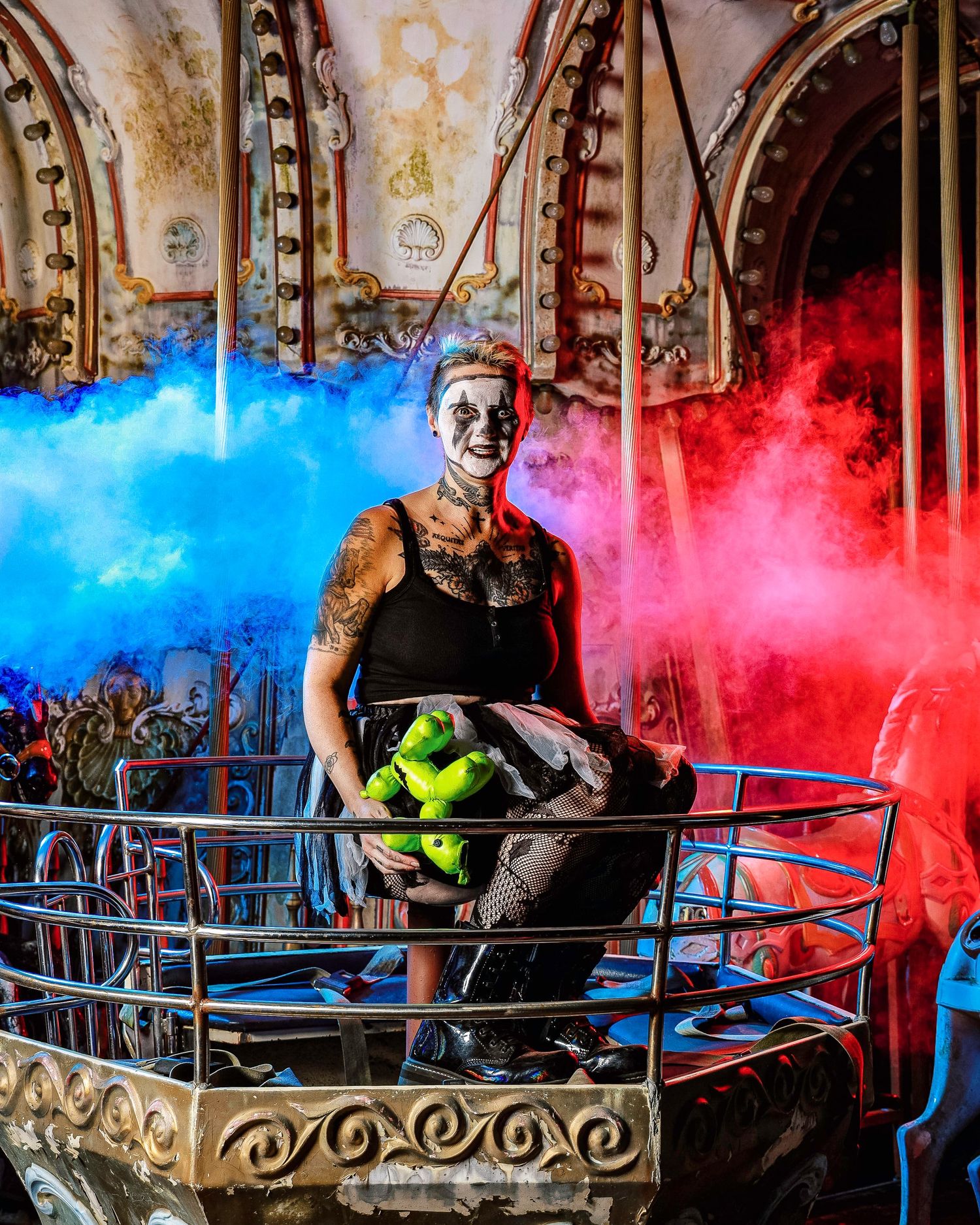 A performer sits in a vintage carnival ride surrounded by colorful smoke effects in blue and red hues.