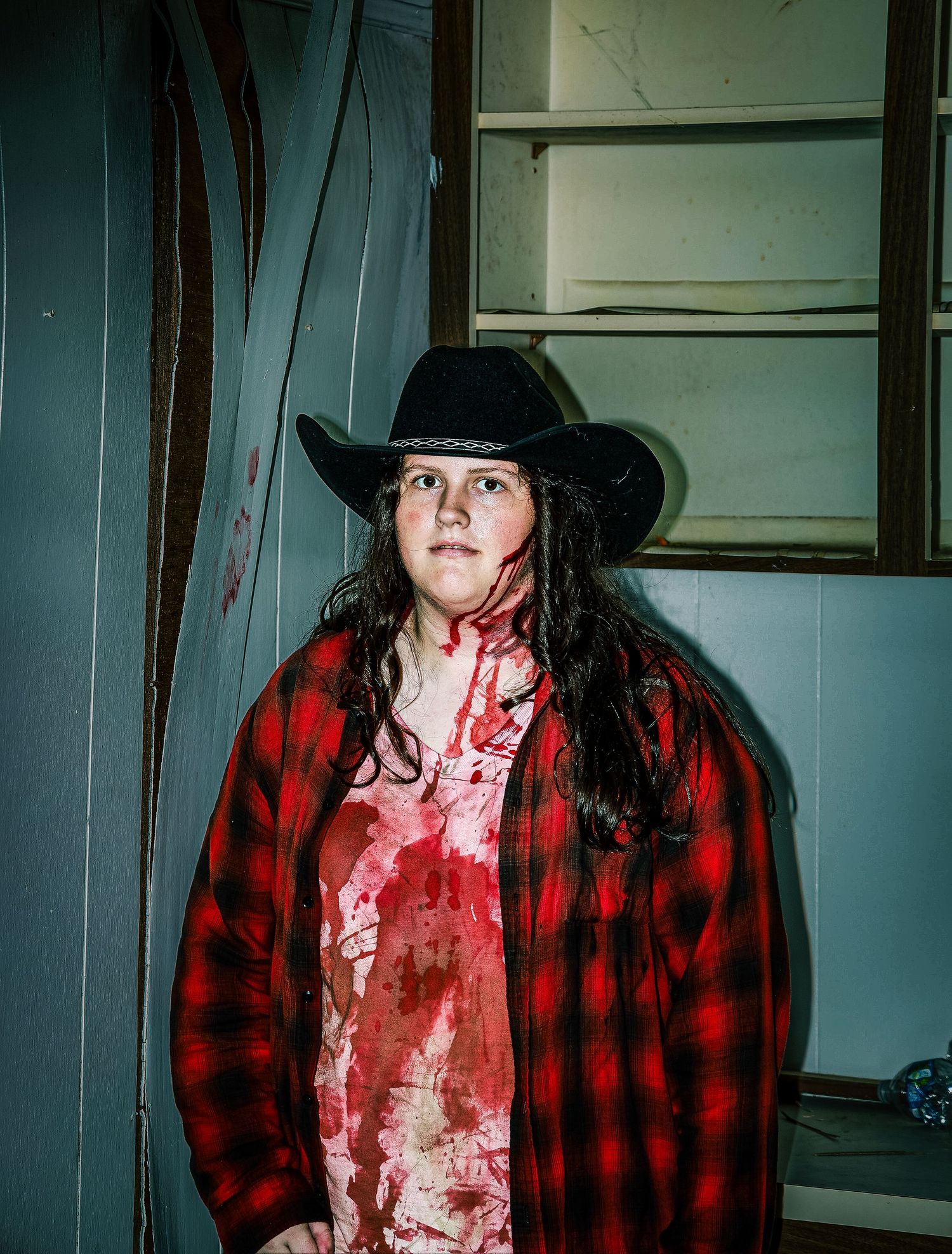 A person in a black hat and red plaid flannel shirt poses in a dimly lit hallway in a horror-themed photo series.