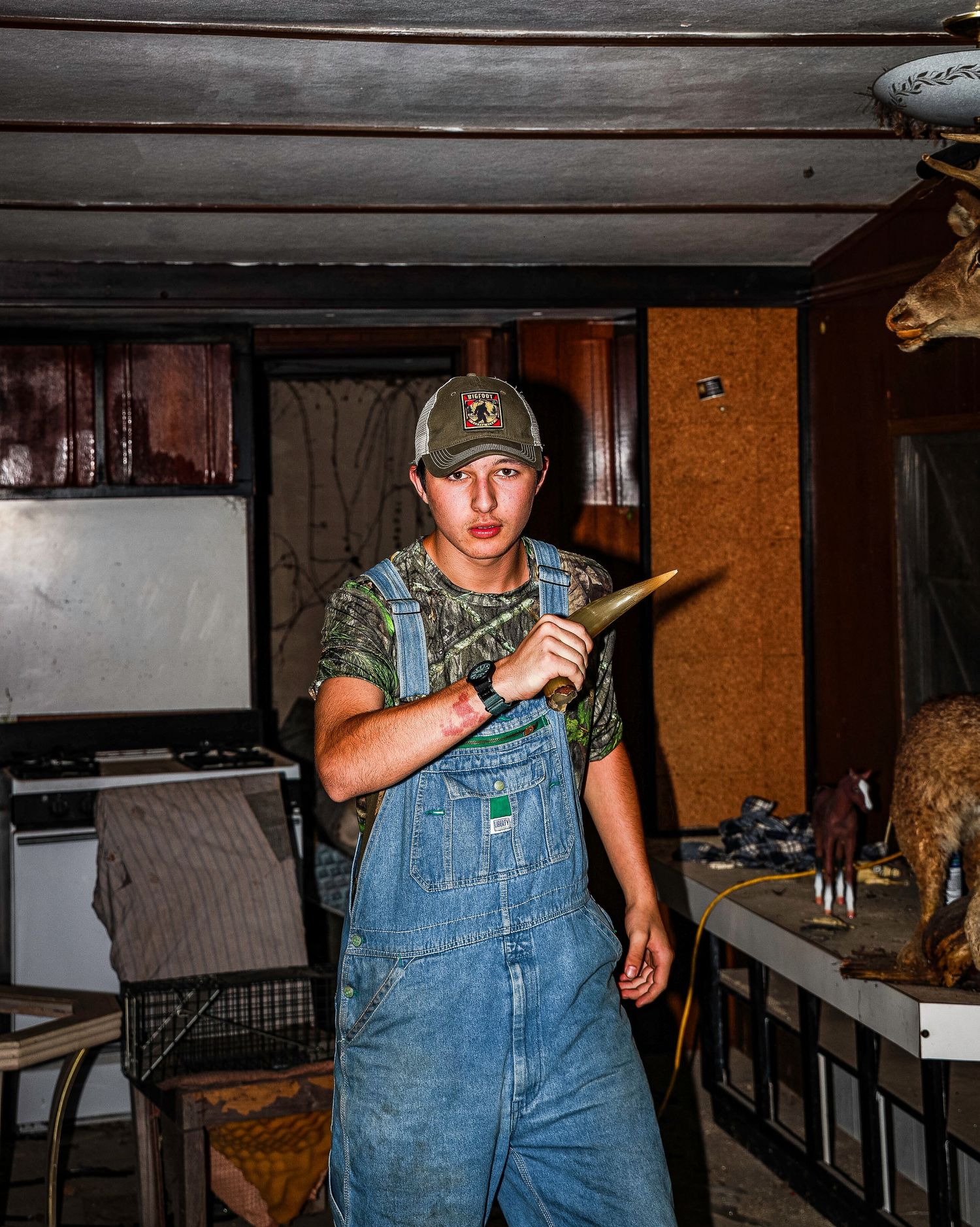 A person in denim overalls stands in a dimly lit room holding a baseball bat in a defensive stance.