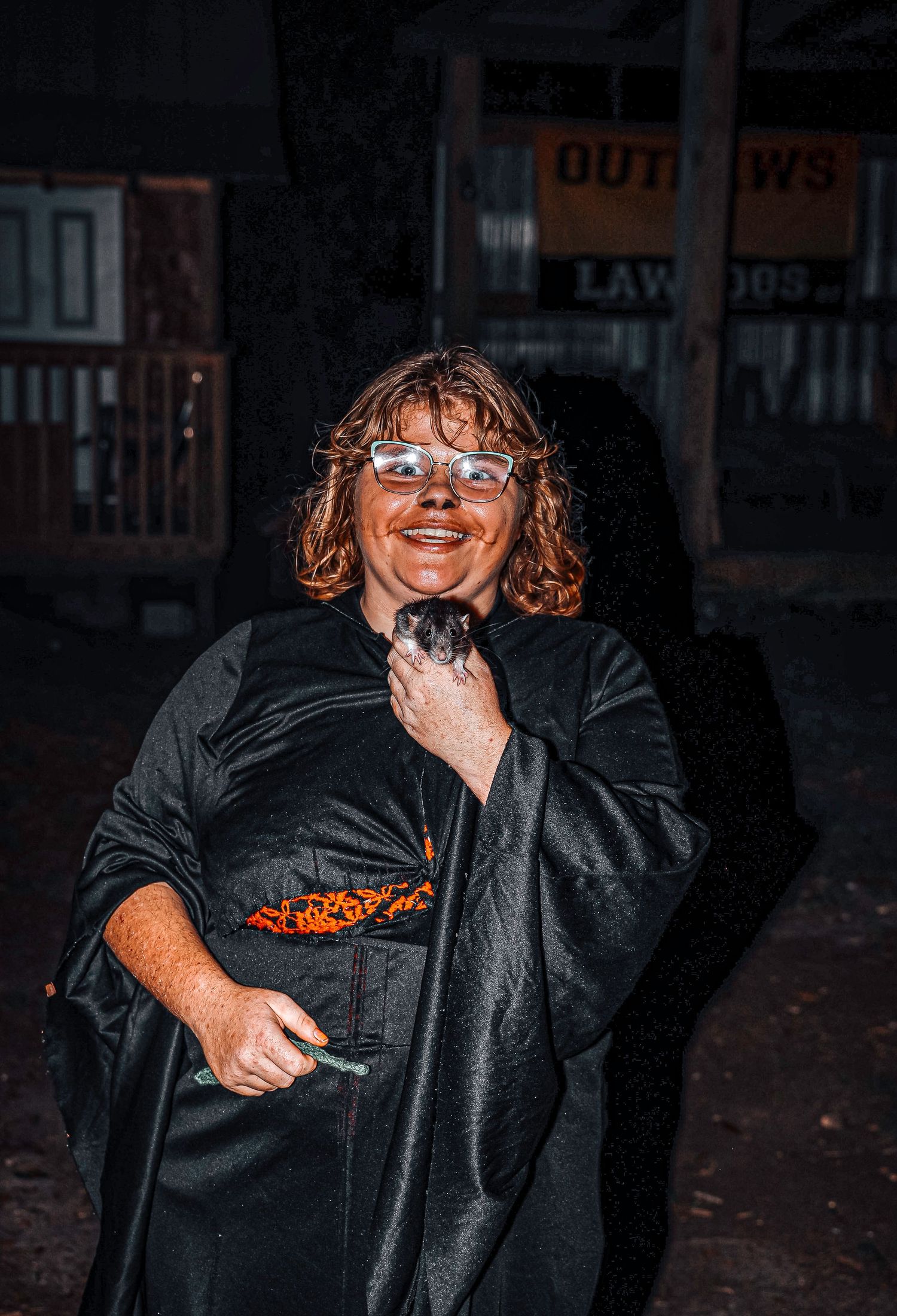 A person wearing a black outfit and glasses smiles joyfully while standing in a dimly lit outdoor setting at night.