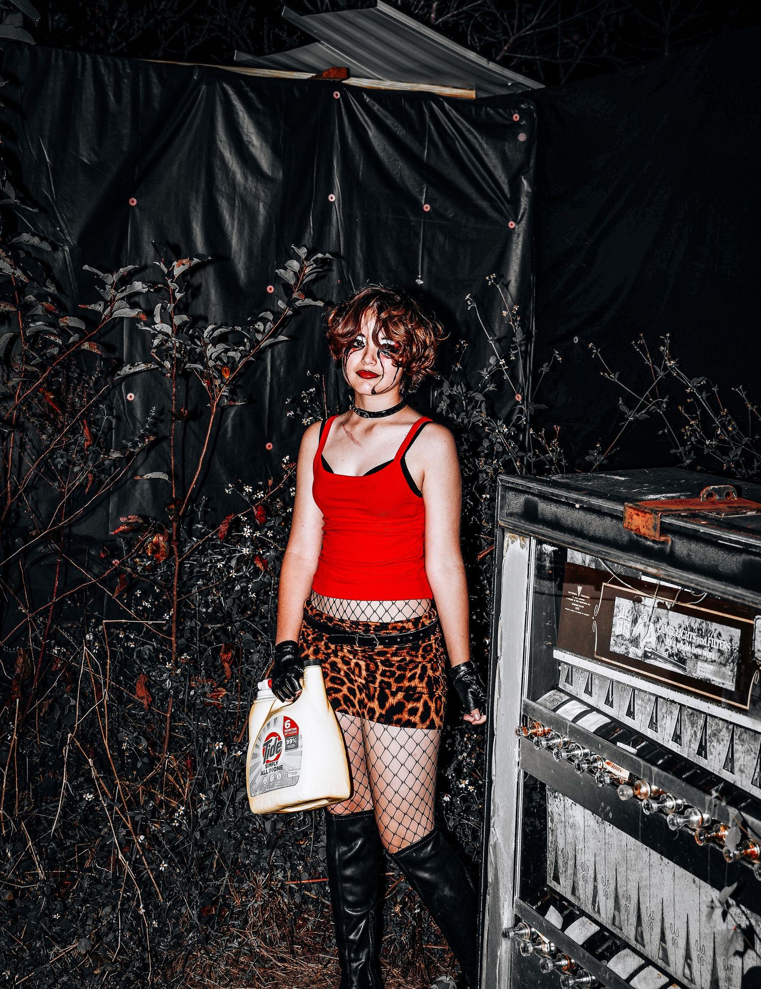 Dark moody scene with someone in a red tank top, leopard print skirt, fishnet stockings and boots holding a white bag.