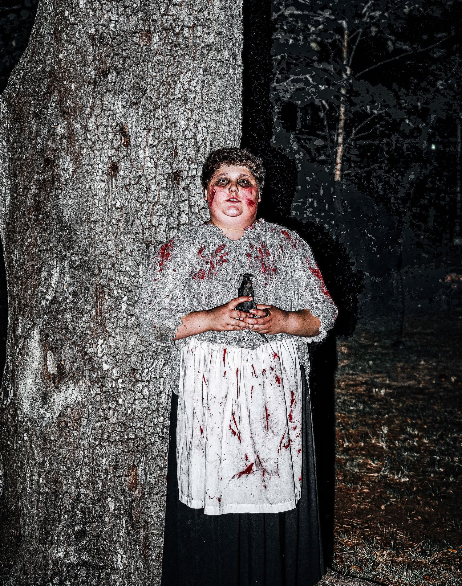 A person in a blood-stained white dress and dark skirt stands against a tree trunk at night in a spooky forest setting.