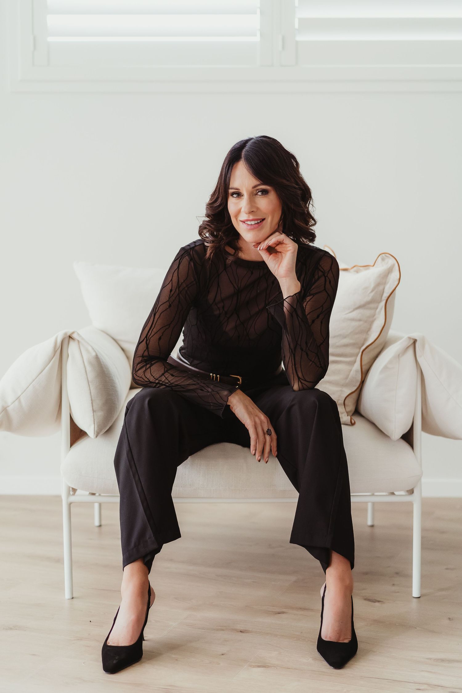 Modern lifestyle portrait of rachael ferguson content creator in a bright setting featuring elegant black outift and heels on a white sofa.