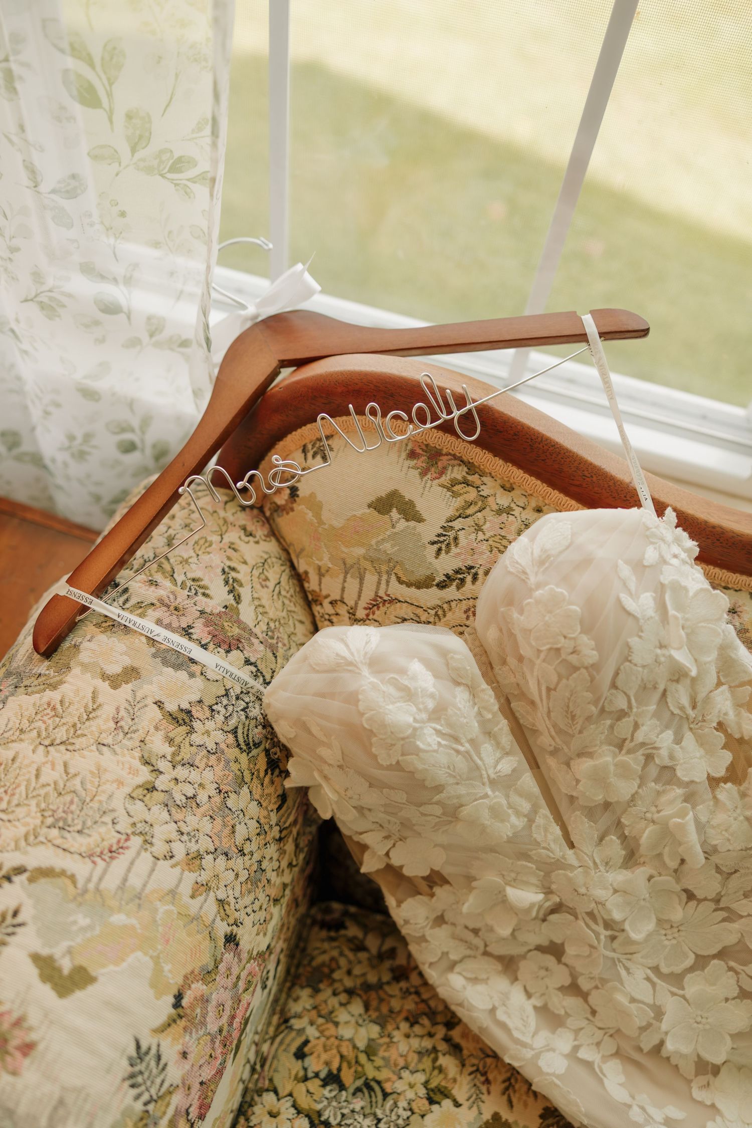 Elegant white lace wedding dress draped over floral chair with personalized wooden hanger near window.
