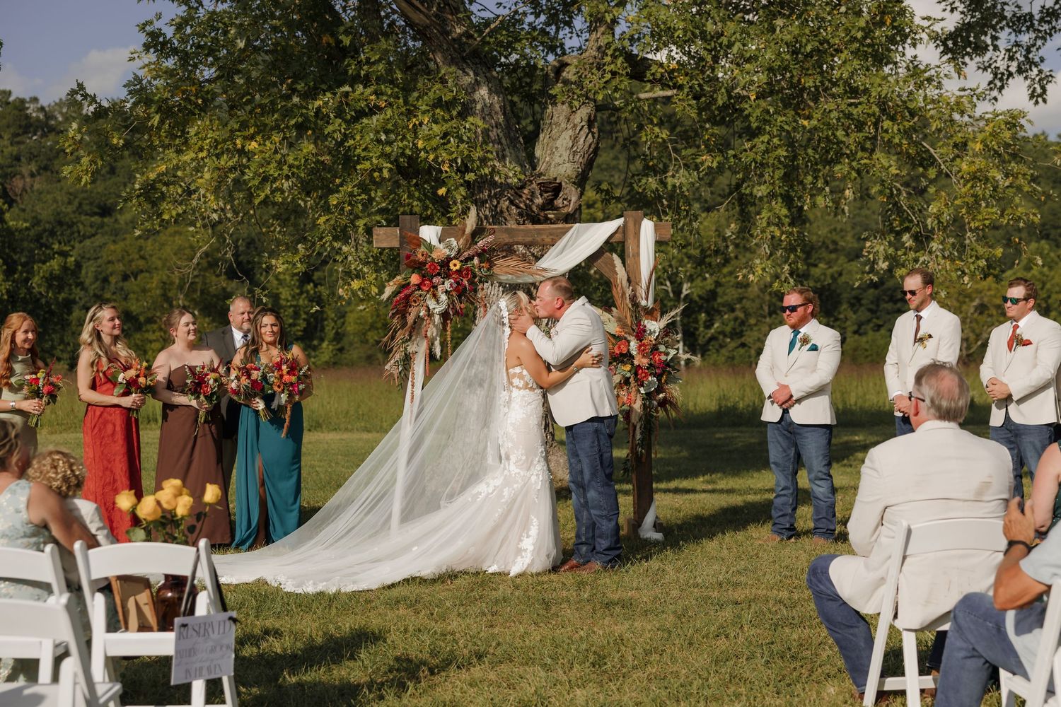 Outdoor wedding ceremony with couple kissing at floral arch, bridal party in colorful dresses and suits on green lawn.