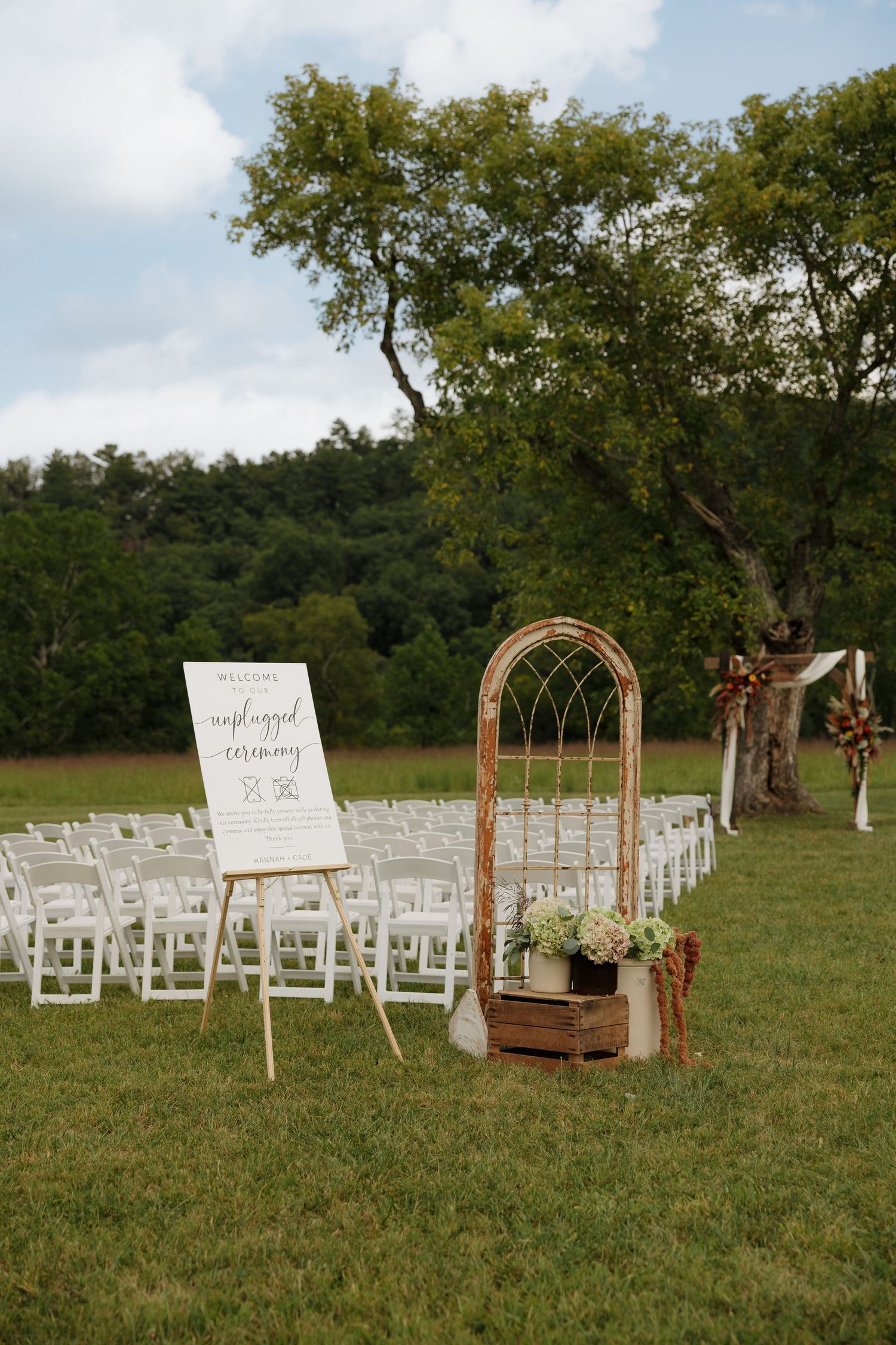 Outdoor wedding ceremony setup with white chairs, rustic arch, welcome sign, and lush green trees in background.