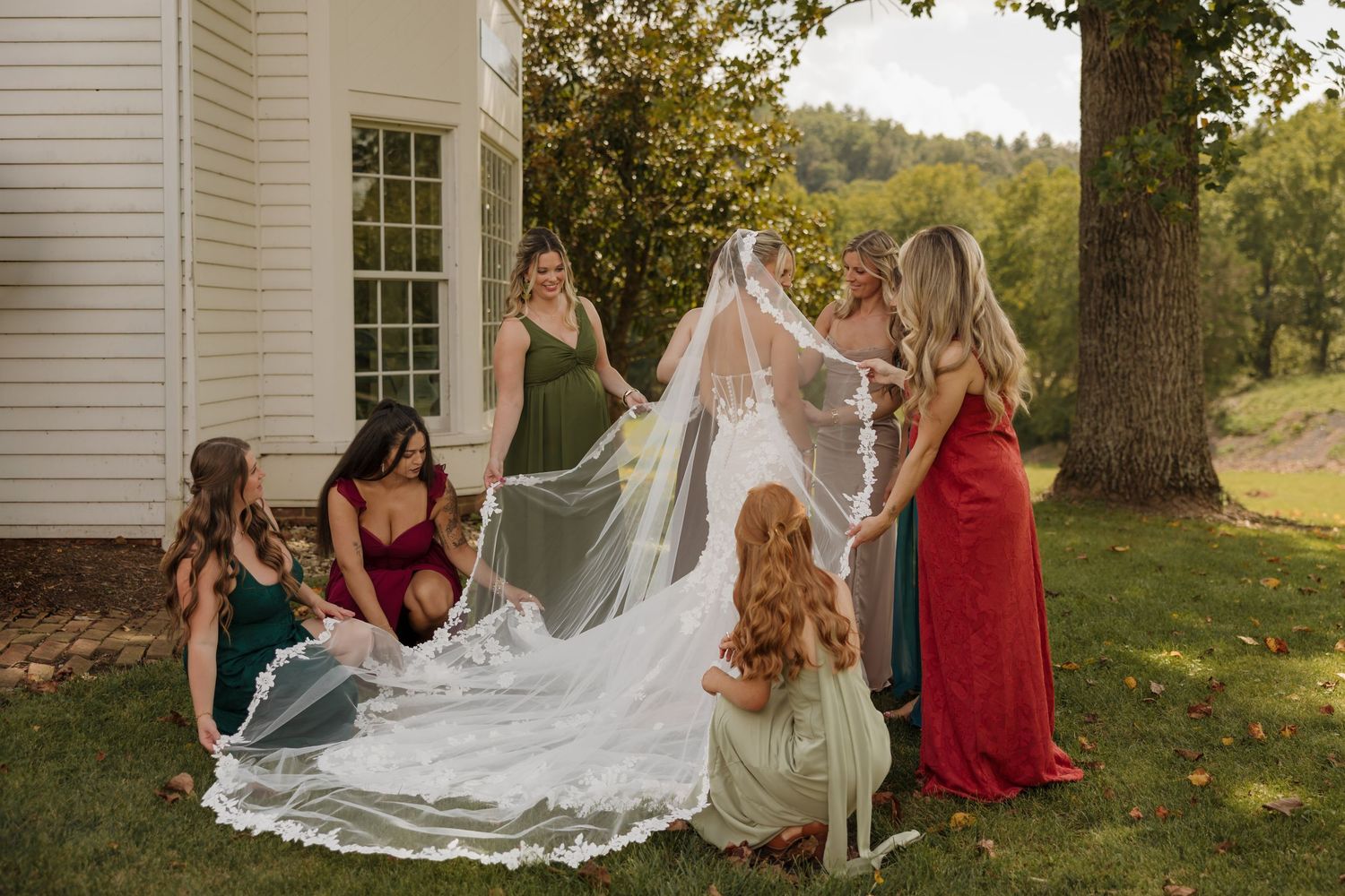 Bridesmaids in colorful gowns gather outdoors to spread a bride's long lace veil near a white building and large tree.