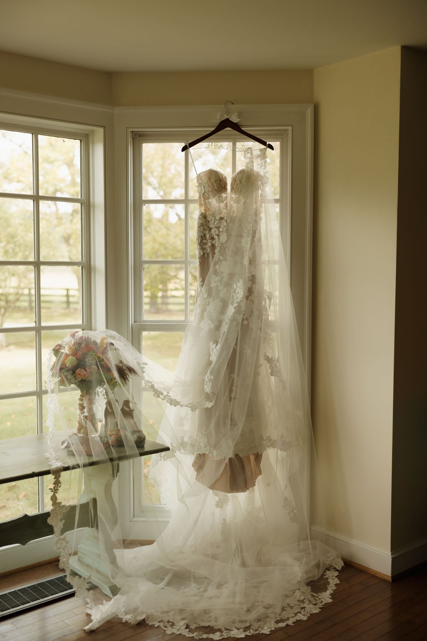 Elegant lace wedding dress hanging in a sunlit bay window with a bridal bouquet and veil displayed nearby.