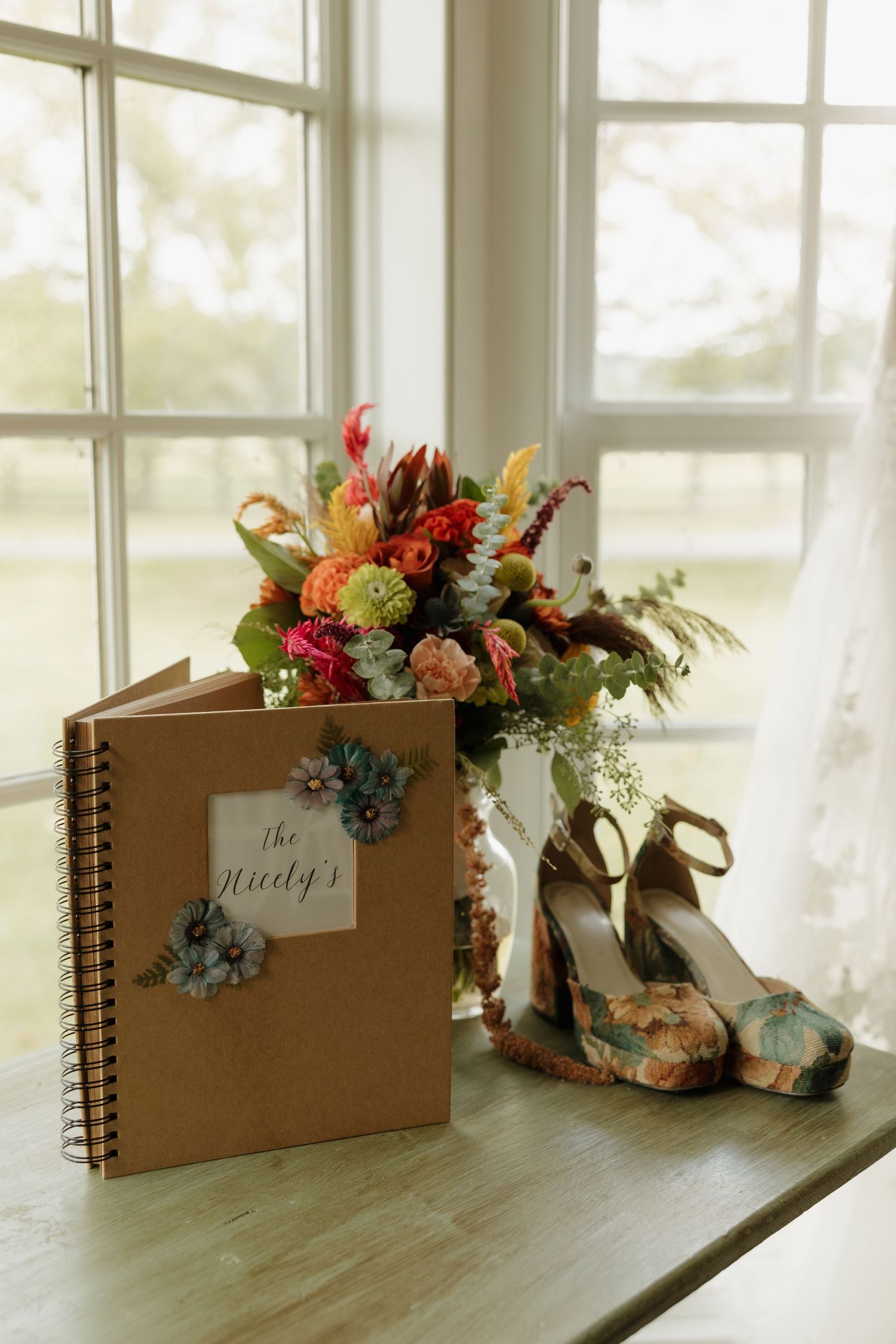 Wedding flat lay featuring a floral bouquet, decorative heels, and a personalized spiral guest book by a sunlit window.