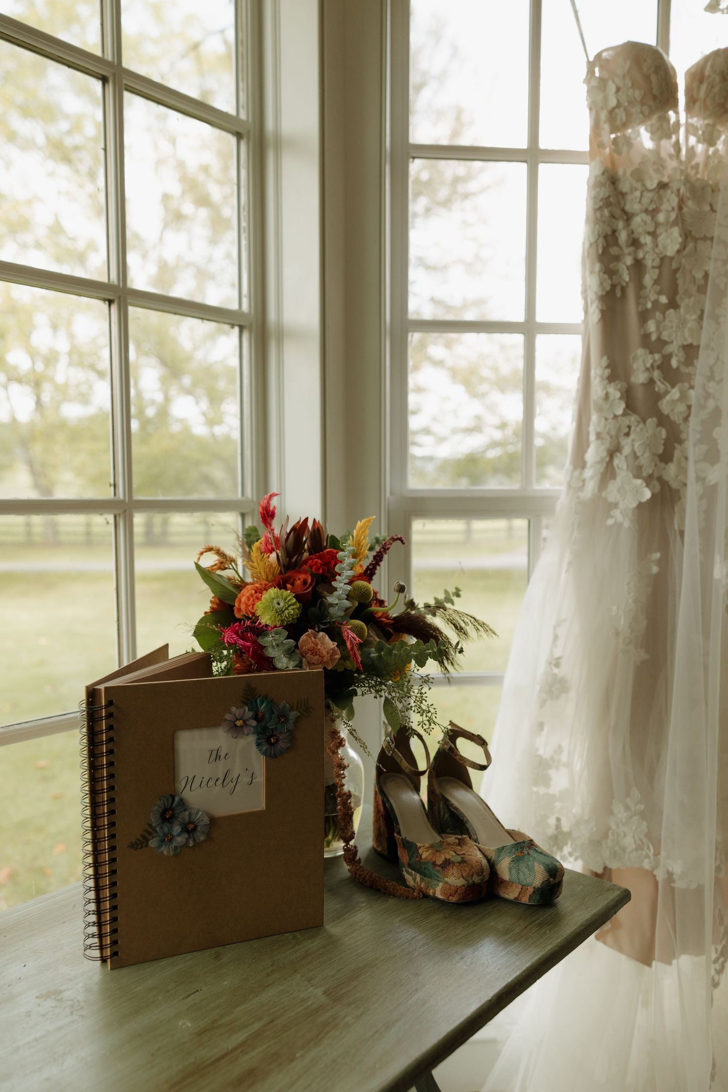 Wedding flat lay featuring a lace bridal gown, colorful bouquet, floral heels, and guest book by a window.