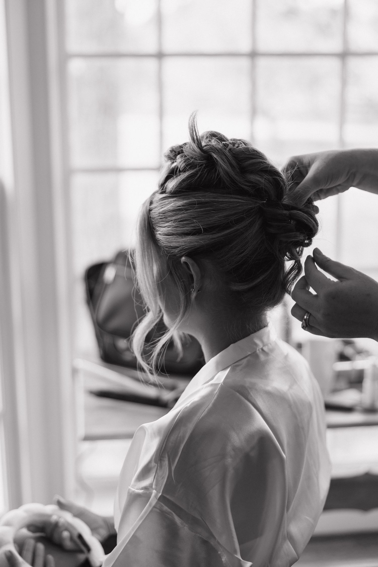 Black and white photo of a bride having her elegant braided updo styled near a bright window on her wedding day.