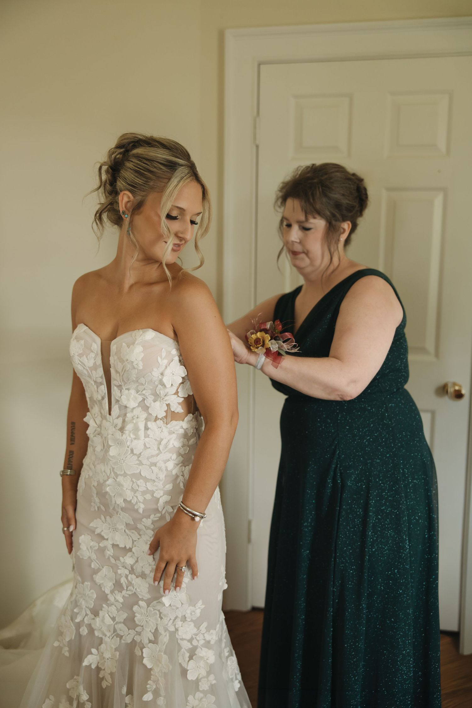 A bride in a floral lace wedding gown being helped by a woman in a dark green dress before the ceremony.