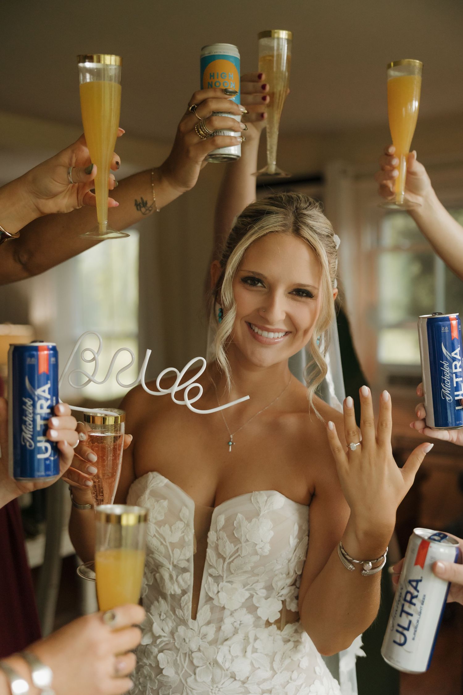 Smiling bride shows off her ring surrounded by friends toasting with mimosas and Michelob Ultra cans.