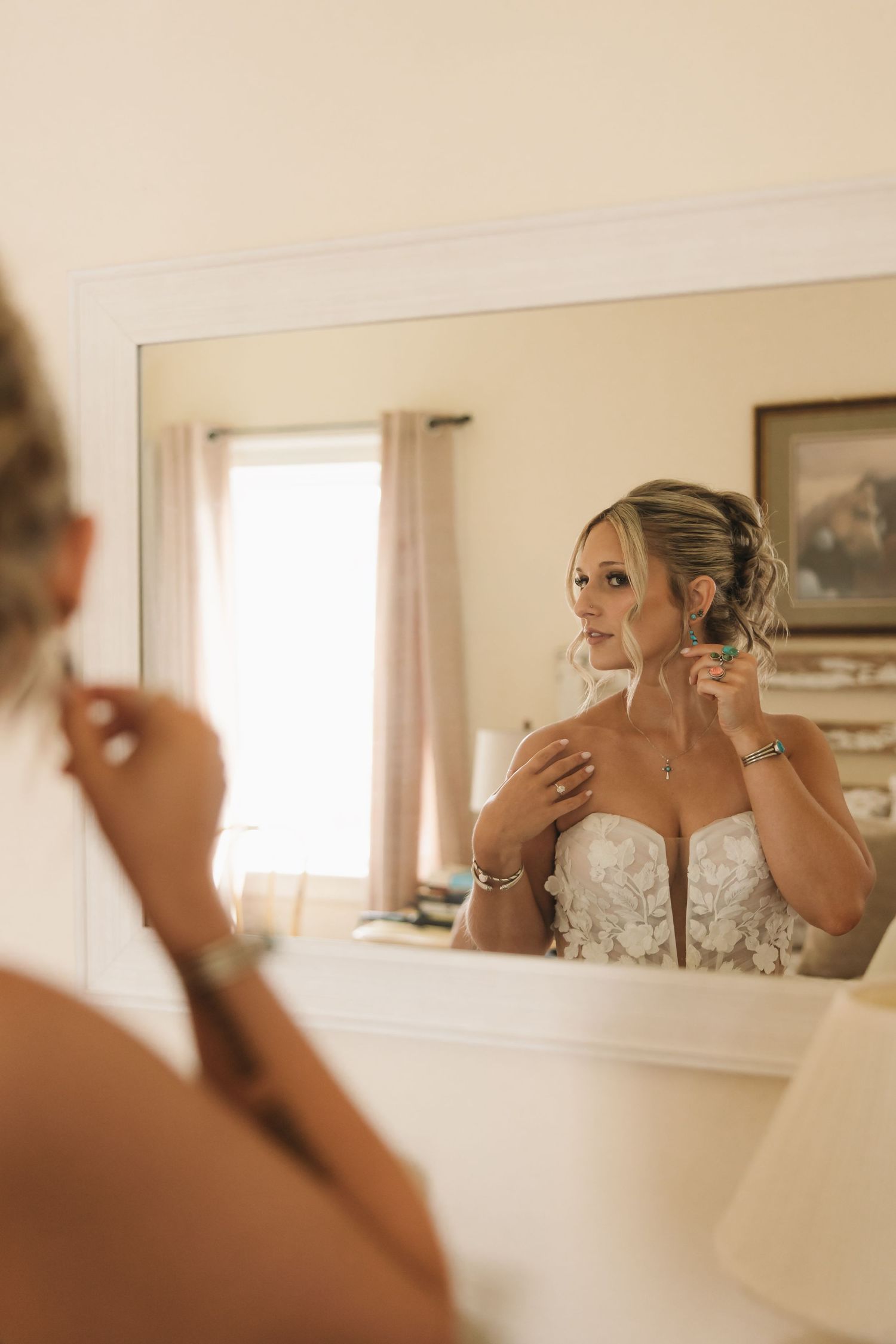 Bride in lace wedding gown putting on earrings while looking at her reflection in a large mirror.
