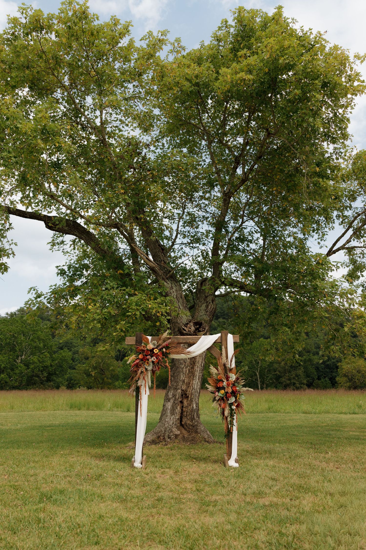 Rustic wooden wedding arch adorned with colorful florals and white draping beneath a large tree outdoors.