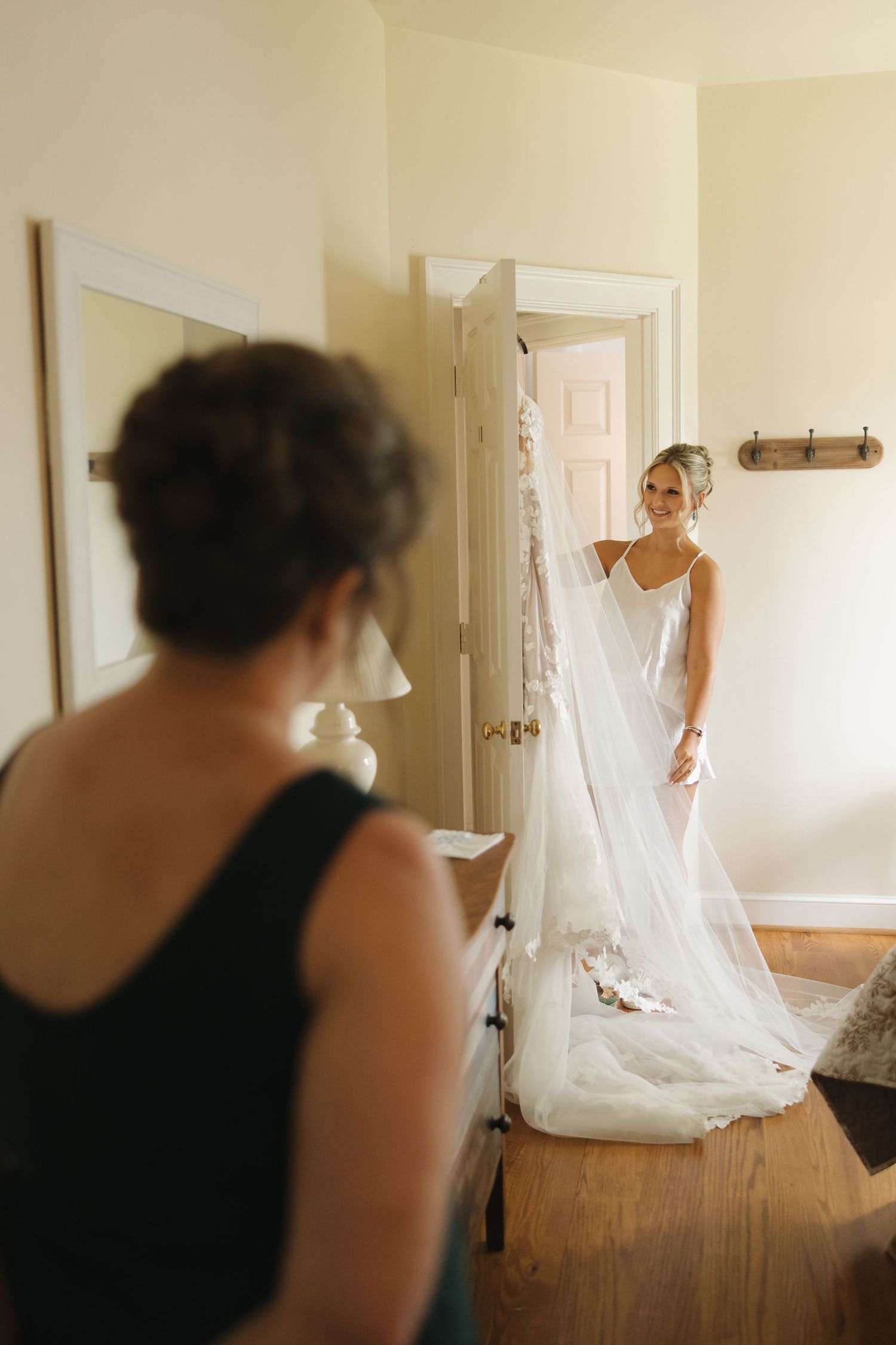 Bride in elegant white gown and veil smiling in doorway while woman watches during wedding prep.