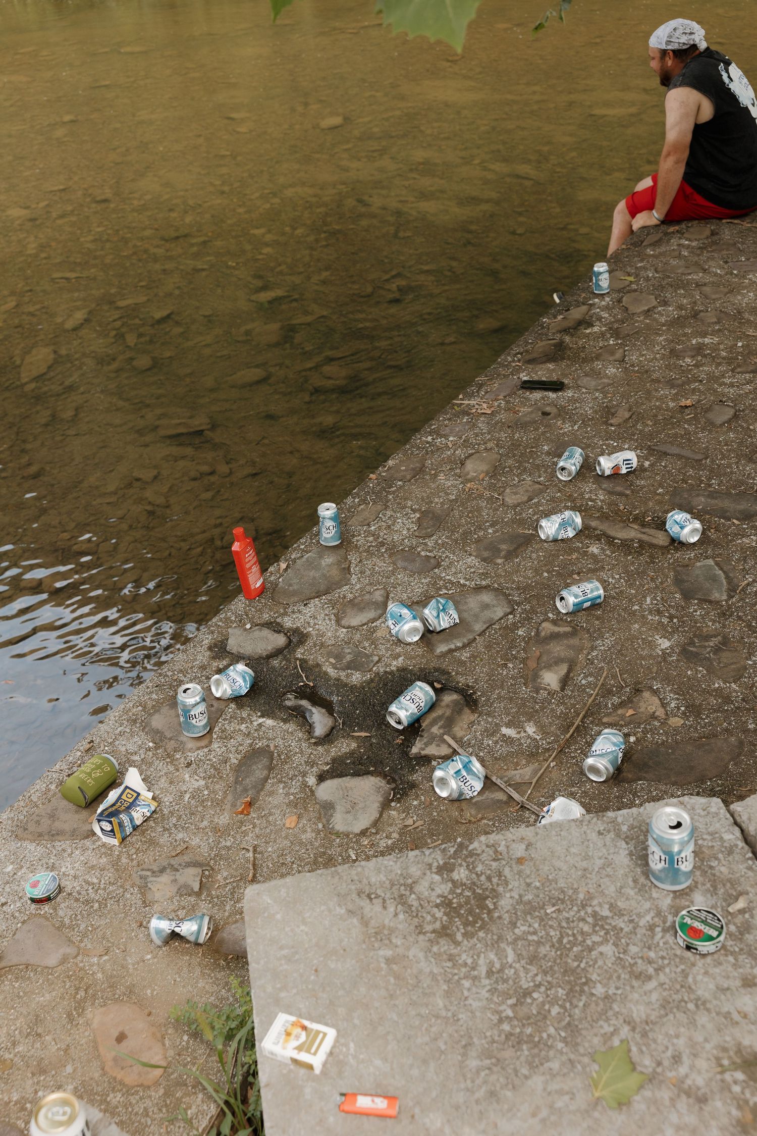 Crushed beer cans and litter scattered on a concrete riverbank ledge near a person sitting by the water.