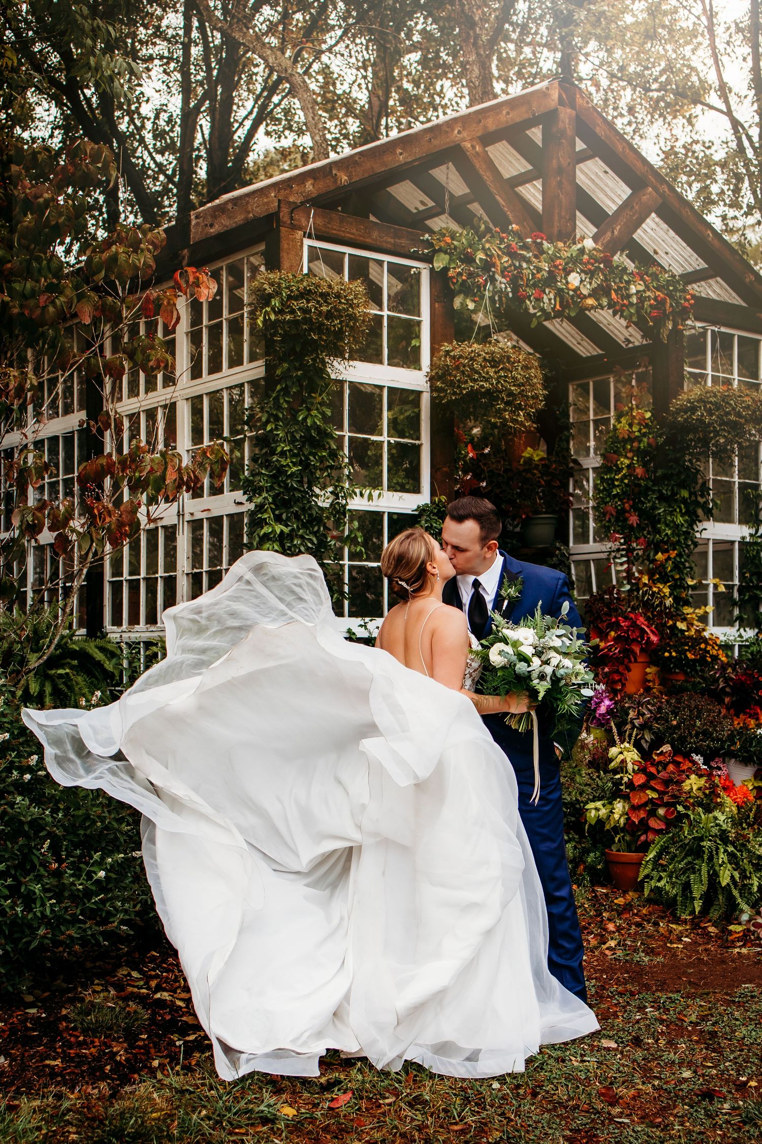 Bride and groom share a kiss outdoors with wedding dress flowing in the wind before a rustic greenhouse backdrop.