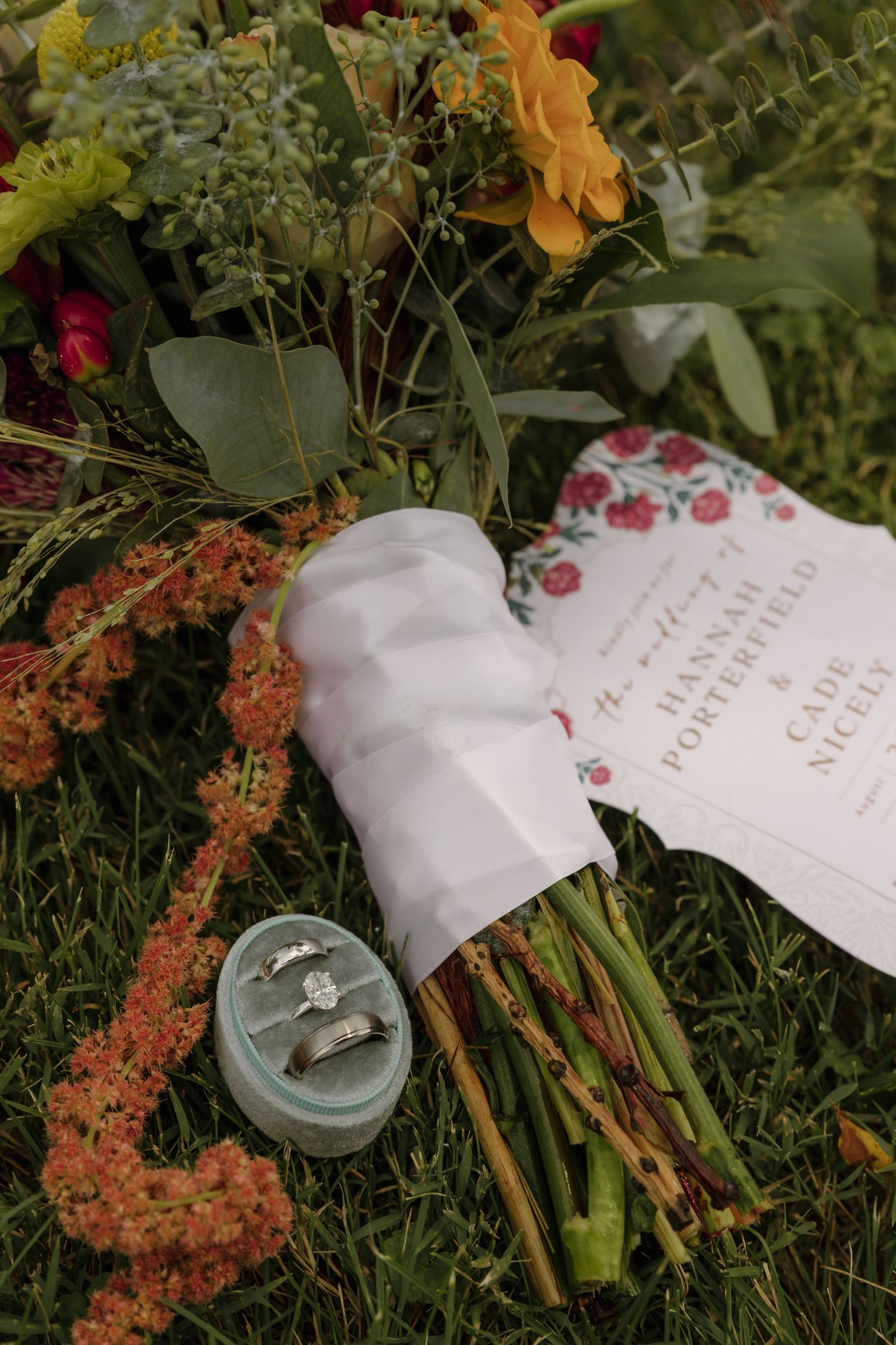 Wedding rings in a teal velvet box beside a bridal bouquet of wildflowers and a floral wedding program on green grass.