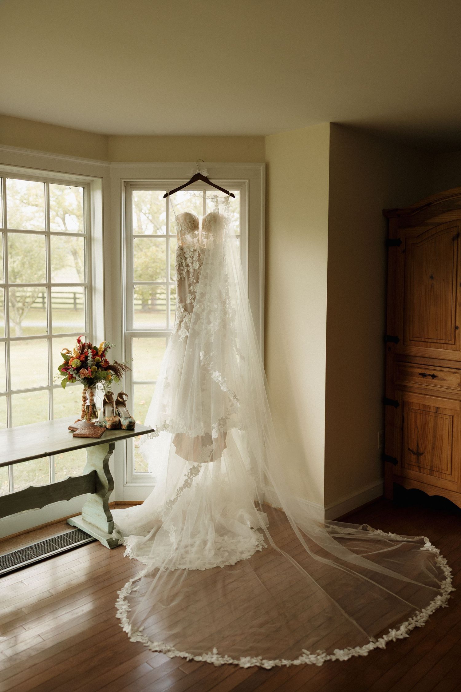 A lace wedding dress with long veil hangs in a sunlit window beside bridal bouquet and heels.