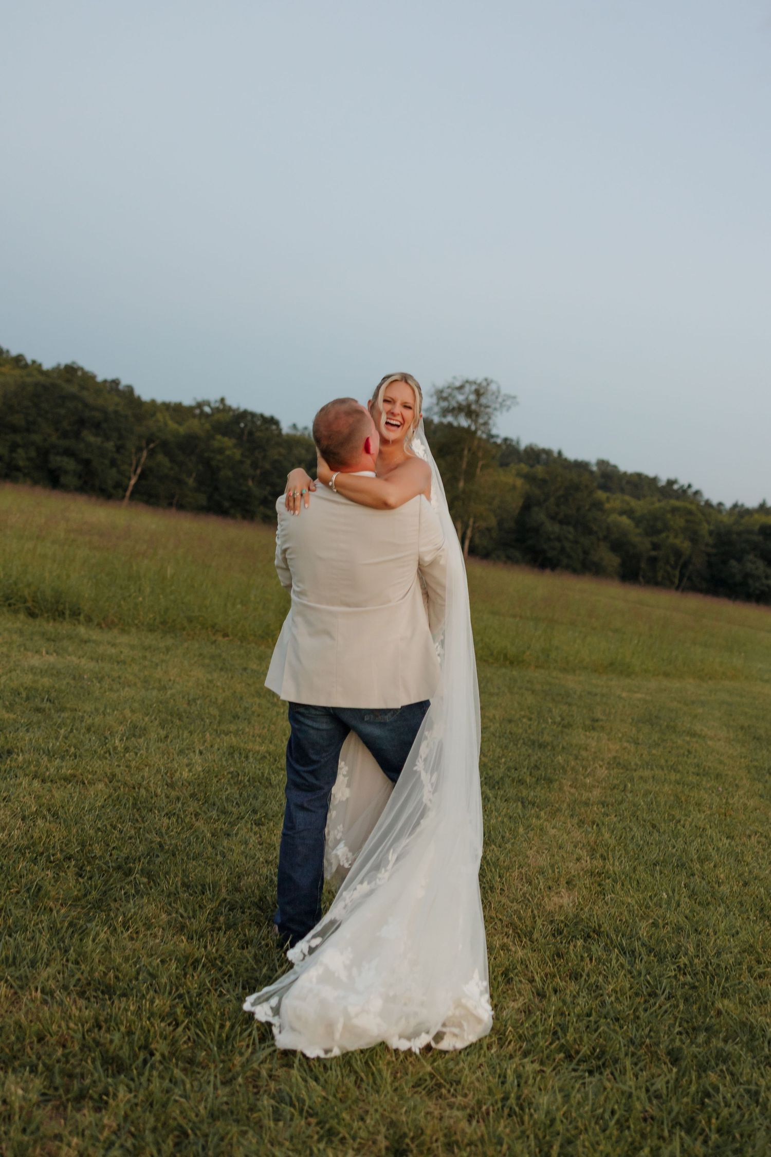 Groom lifting bride in white wedding gown and veil in a lush green field at golden hour.
