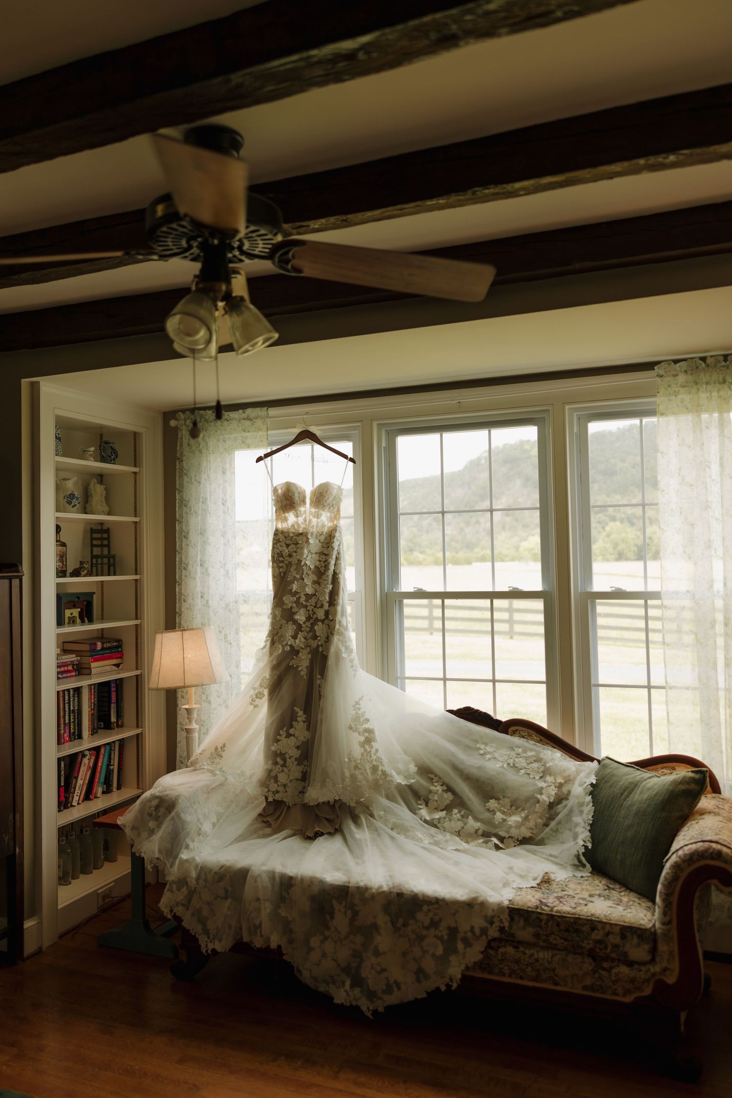 Elegant lace wedding dress hanging on a hanger over a vintage floral sofa in front of large windows in a cozy room.