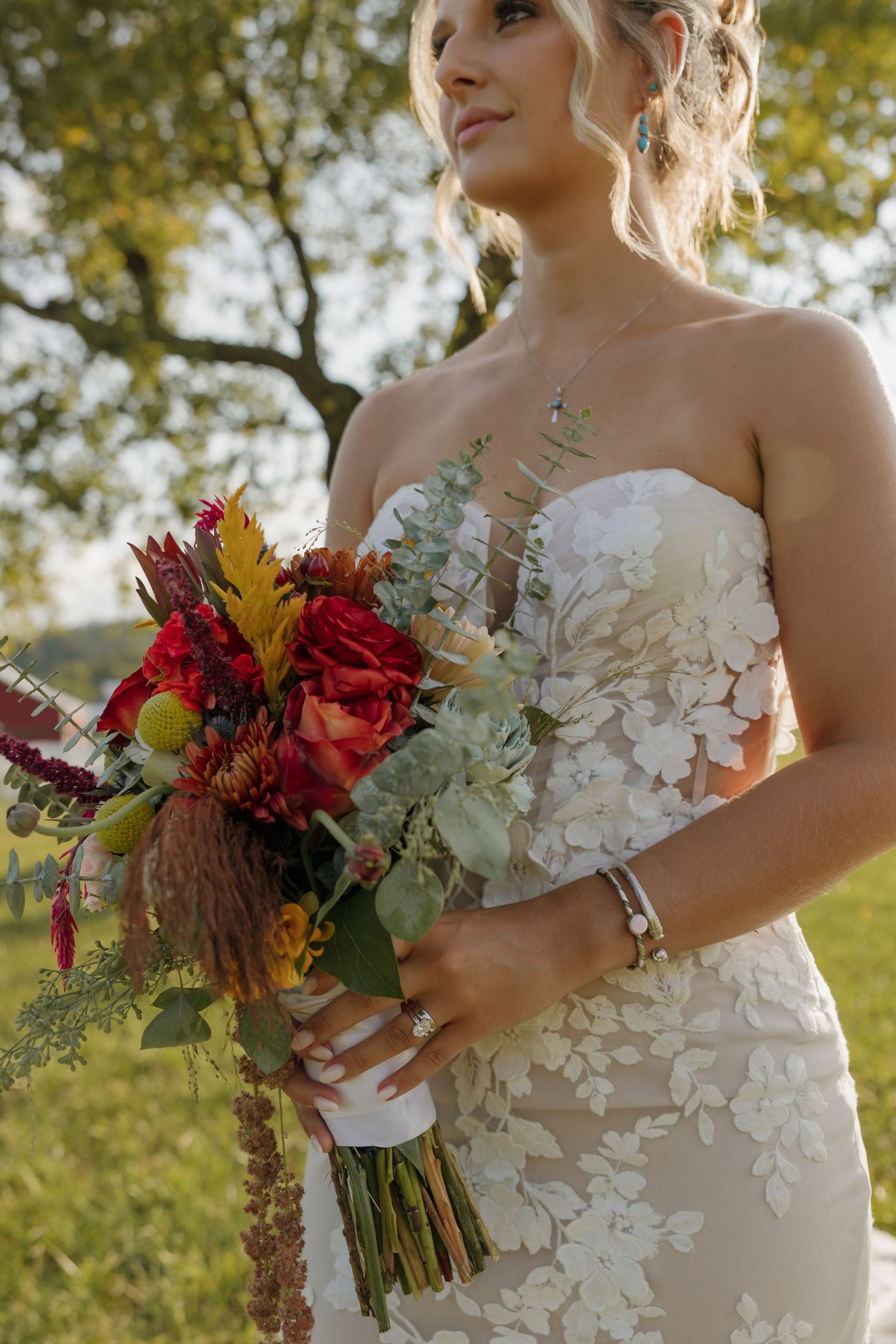 Bride in strapless lace gown holds vibrant red and orange bouquet with eucalyptus outdoors.