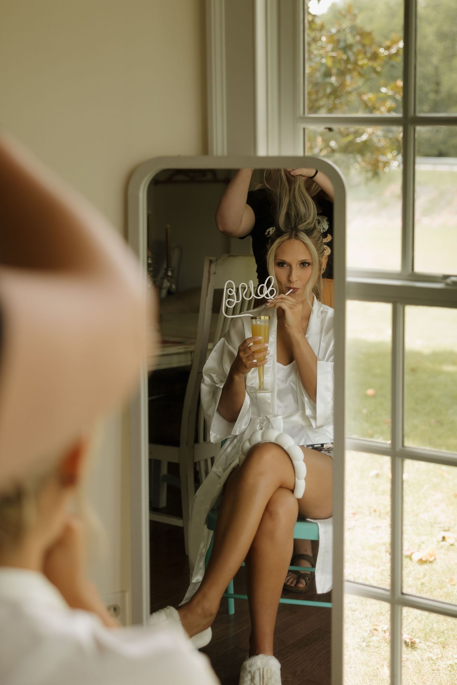 Bride in white robe sips a mimosa while getting ready, reflected in a mirror by a bright window.