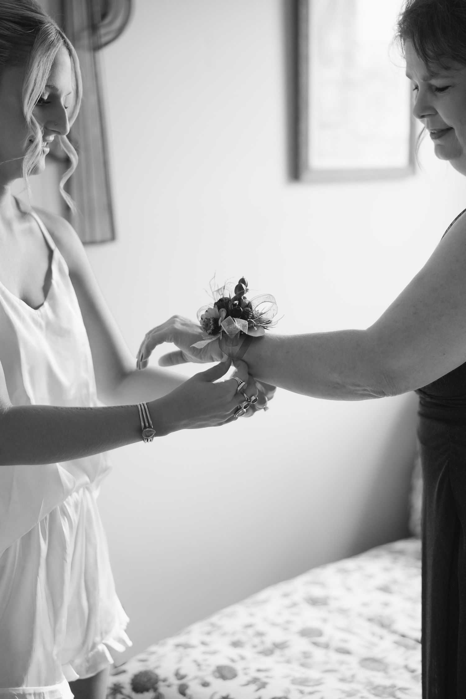 Black and white photo of a bride having a floral wrist corsage placed on her wrist by a woman in a dark dress.