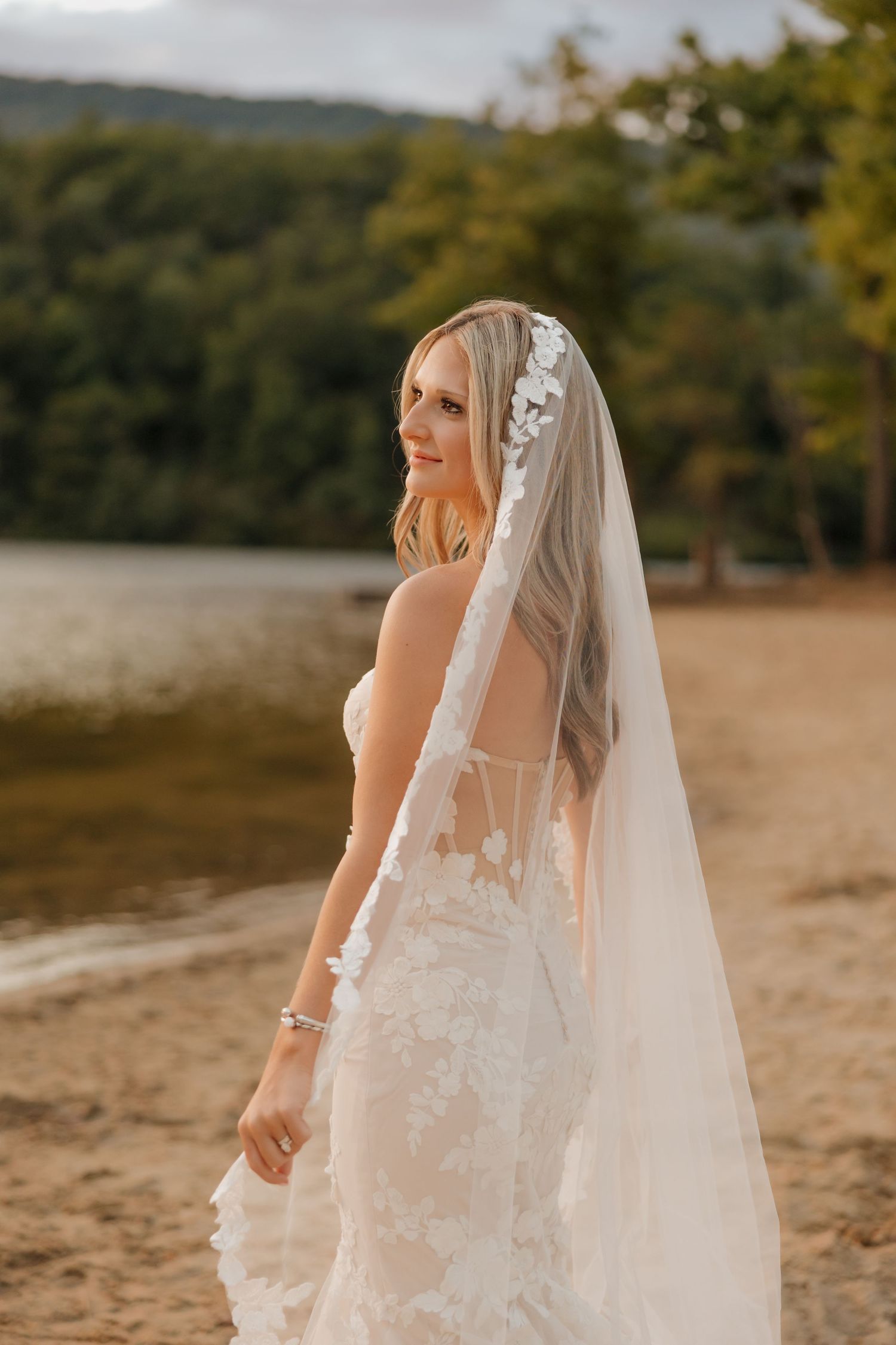 Bride in lace wedding dress and floral veil smiling by a lake with green trees in background.