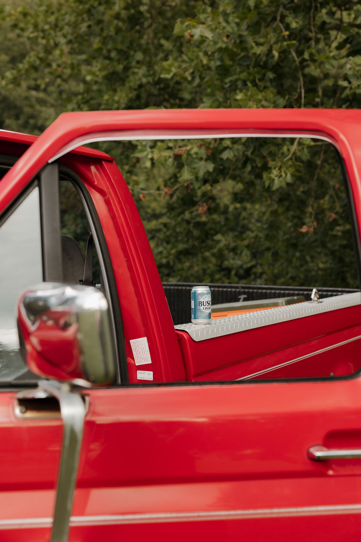 Red pickup truck with open rear sliding window and a beverage can resting on the truck bed rail, parked near green trees.