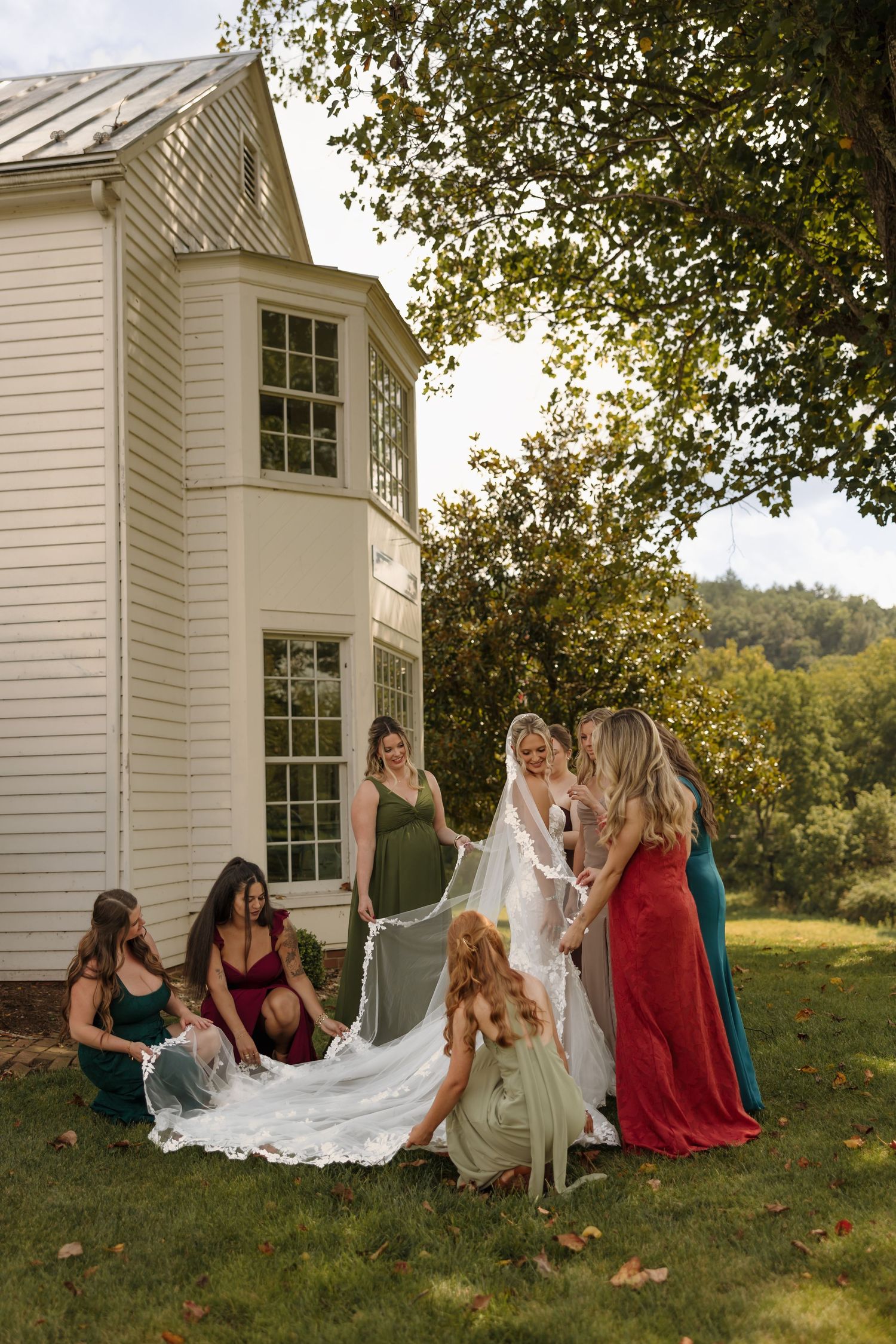 Bridesmaids in colorful gowns arrange a bride's flowing veil on green lawn beside a white country house.